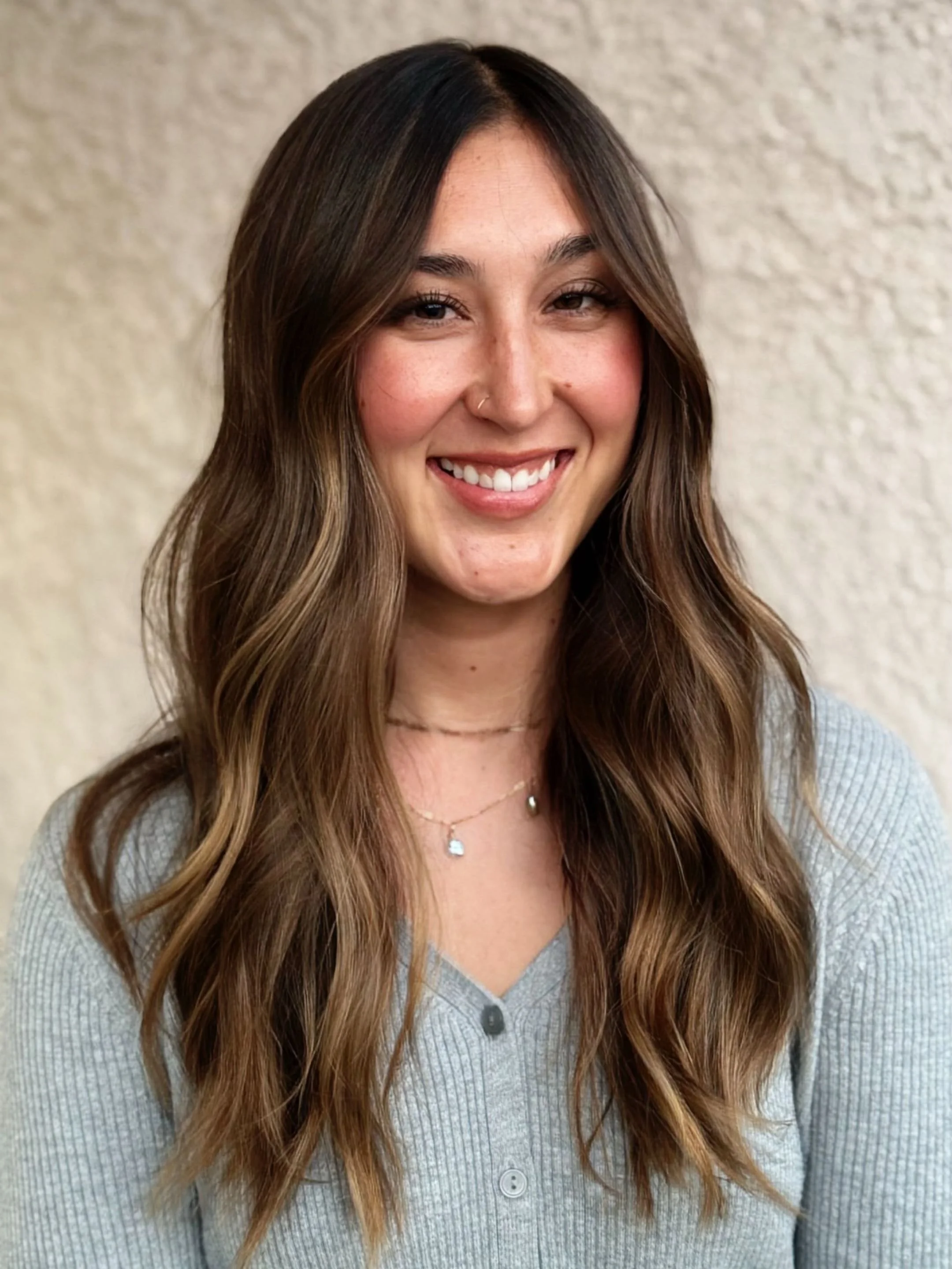 A young woman with long, wavy brown hair and a smile, wearing a light gray cardigan and layered necklaces against a neutral textured background.