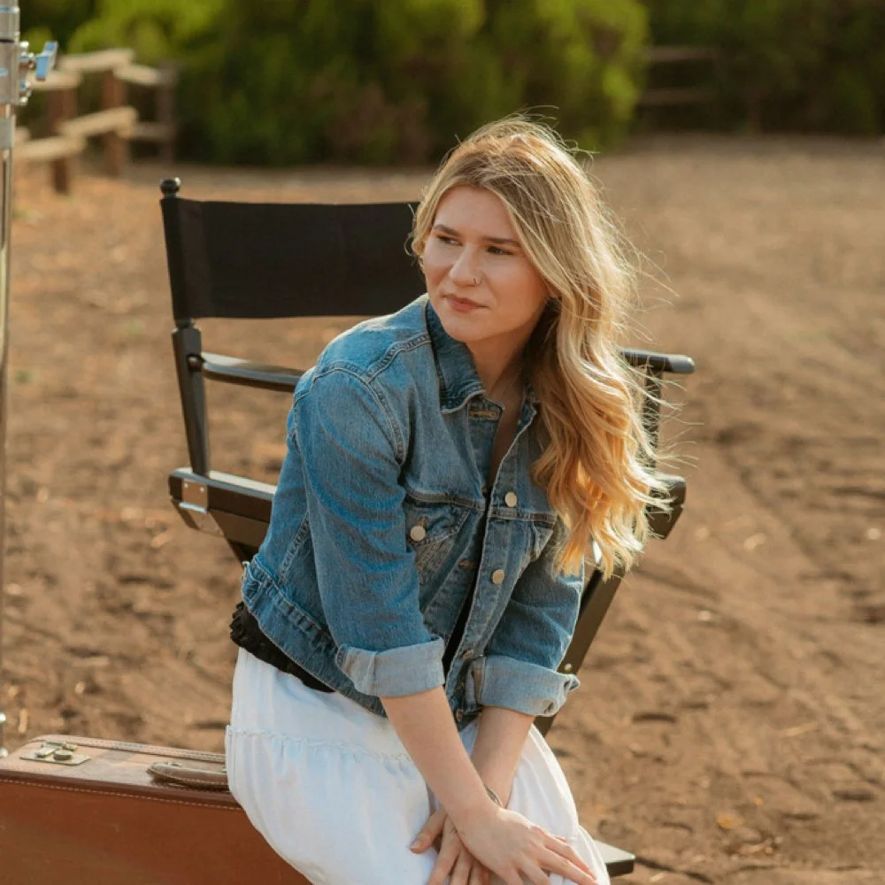 A young woman with wavy blonde hair, wearing a denim jacket and a white dress, sitting outdoors against a black director's chair, with a suitcase beside her, on a dirt path surrounded by greenery.