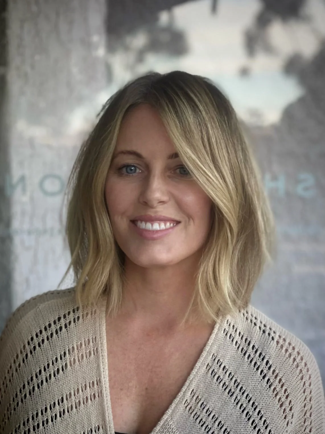 A smiling woman with blonde, shoulder-length wavy hair, wearing a beige knit top, standing in front of a textured background.