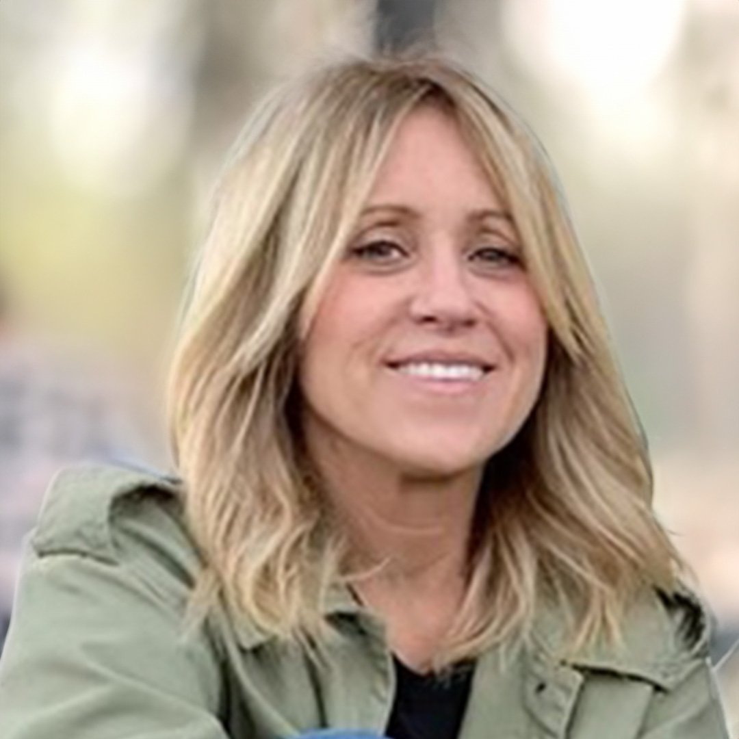 A woman with shoulder-length blonde hair smiling outdoors in a green jacket.
