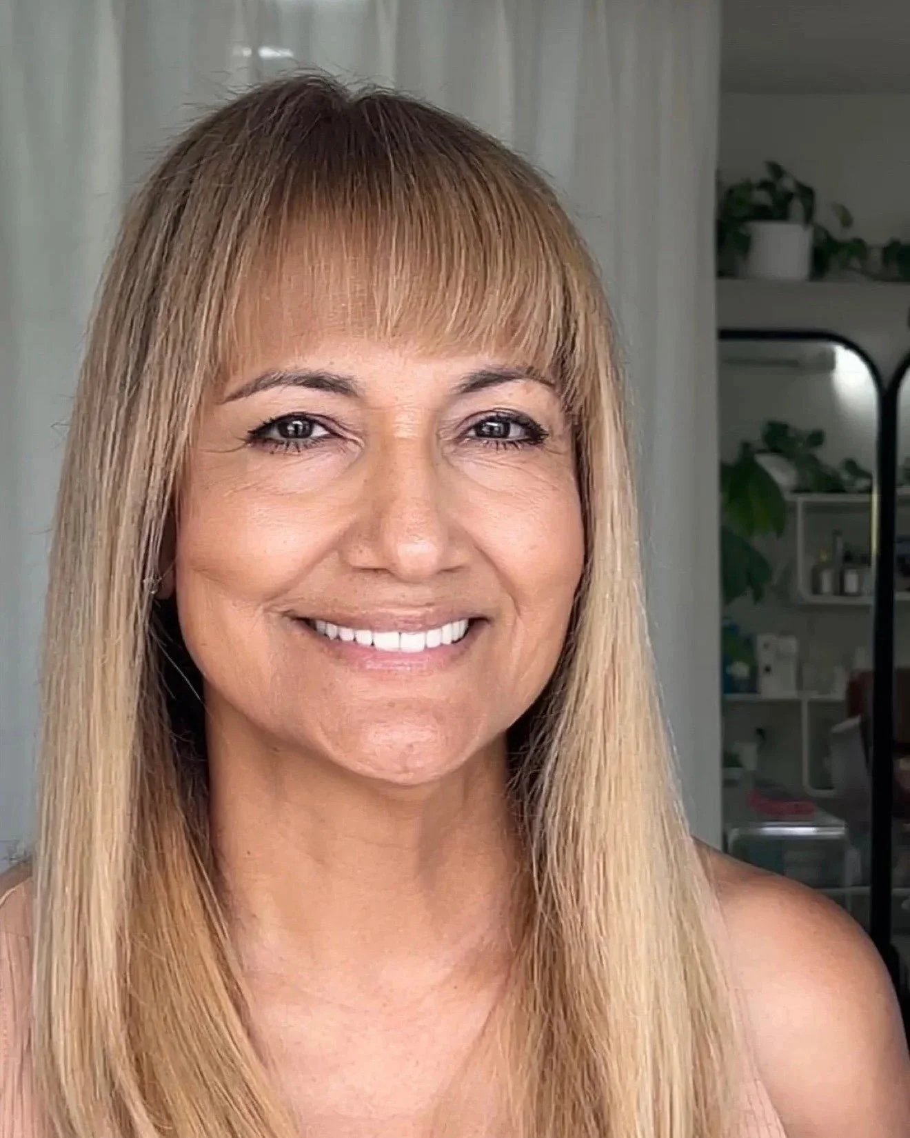 A smiling woman with long blonde hair and bangs, wearing natural makeup. In the background, there are shelves with plants and household items.