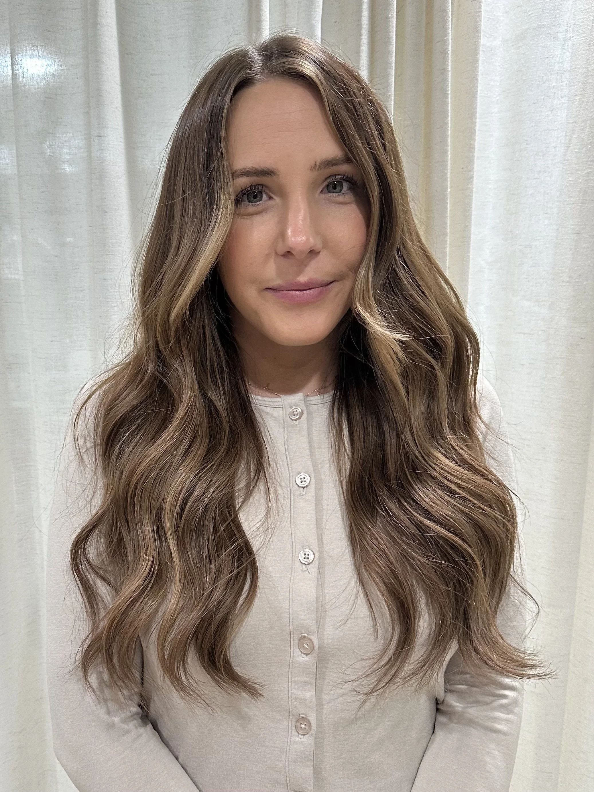 A woman with long, wavy brown hair, wearing a white button-up shirt, standing in front of a light-colored curtain.