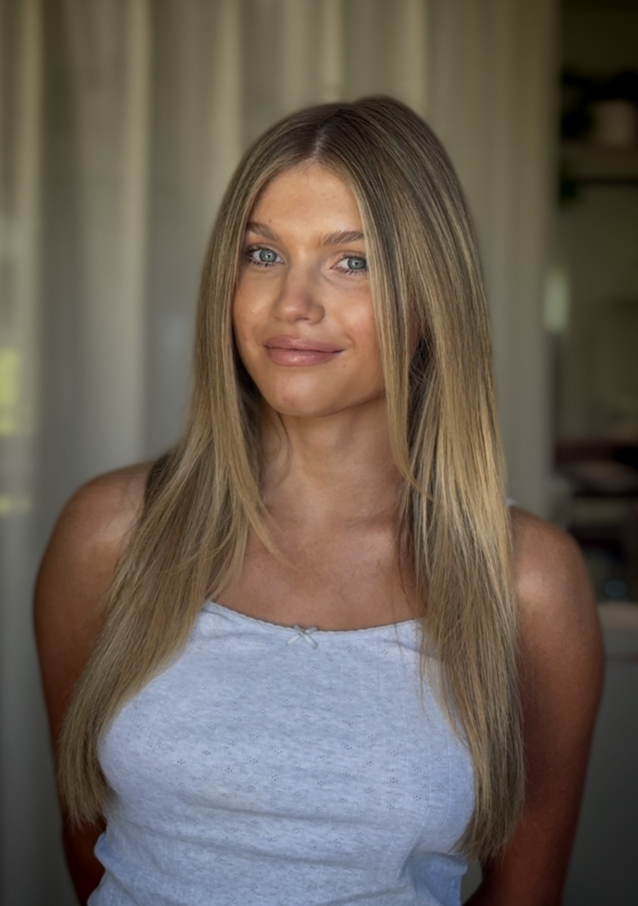 A woman with long, straight, blonde hair and blue eyes, smiling, wearing a white tank top, in a room with curtains and shelves in the background.