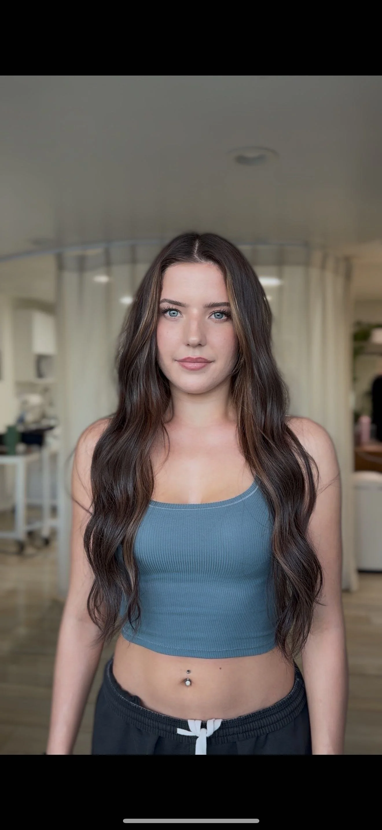 Young woman with long wavy brown hair and blue eyes standing indoors, wearing a blue crop top and dark pants.