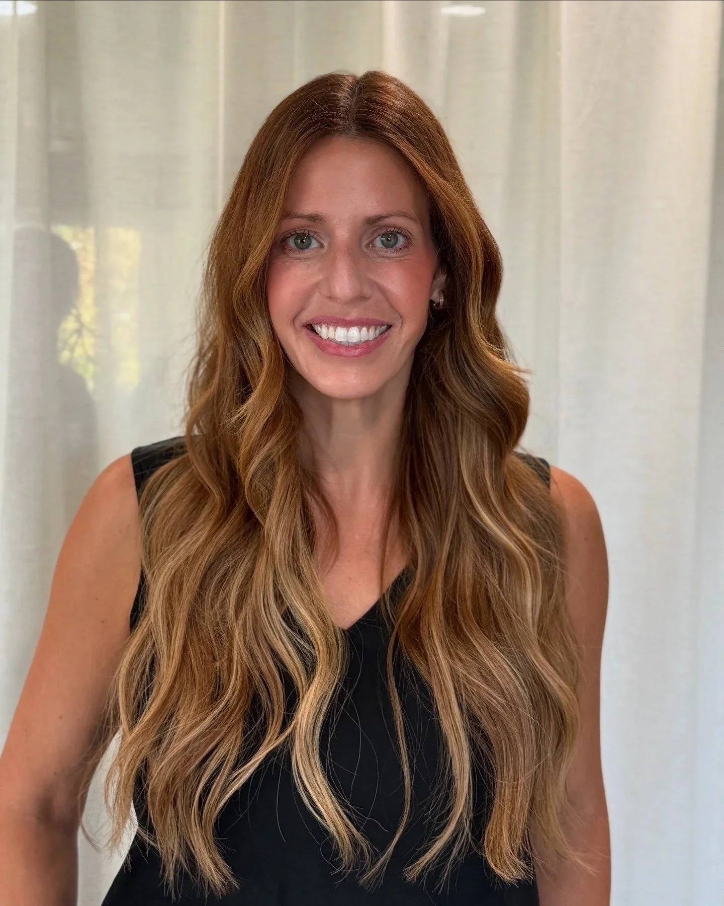 Portrait of a woman with long wavy blonde hair wearing a black sleeveless top, smiling at the camera, with light-colored curtains in the background.
