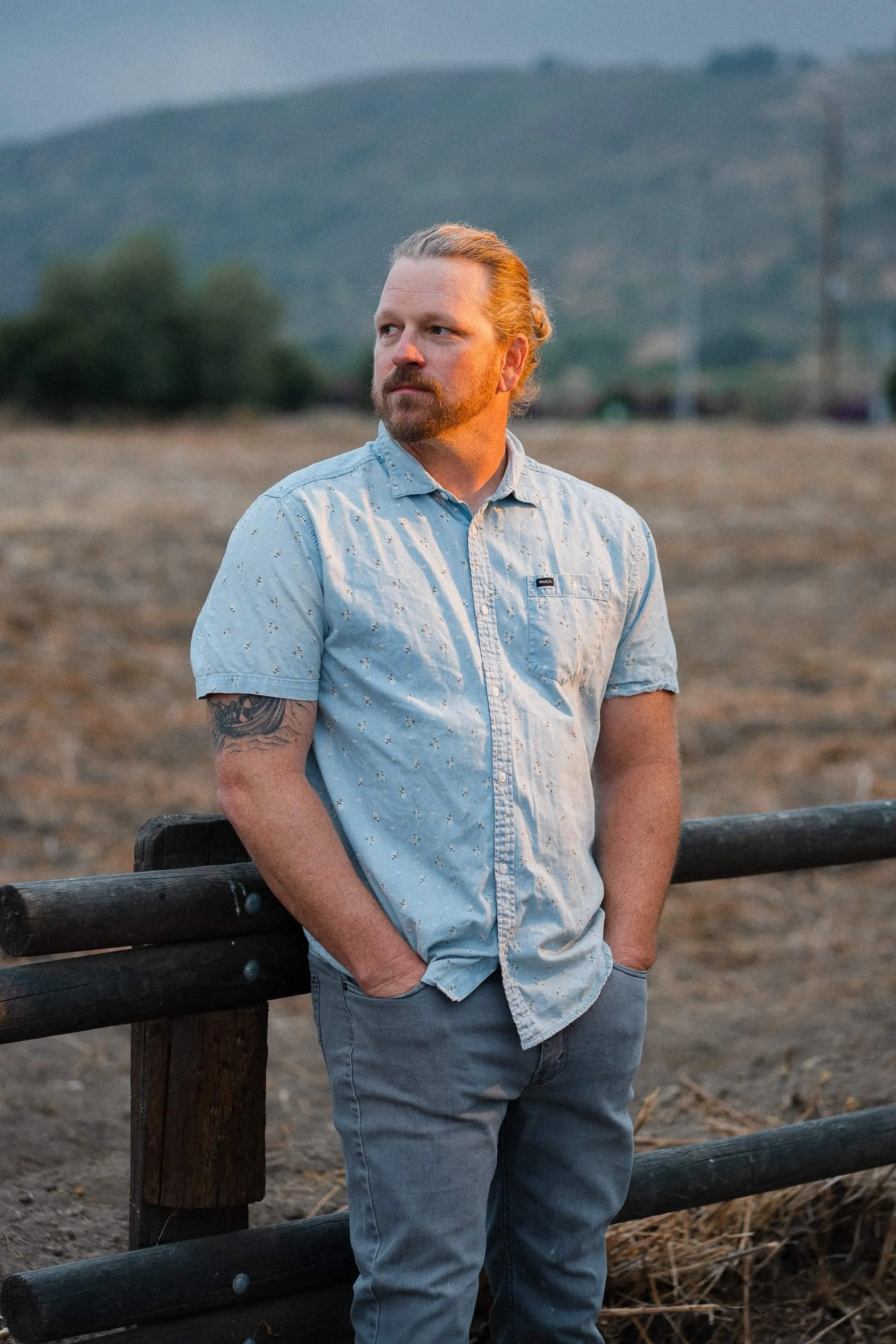 A man with a beard and long hair tied back, wearing a light blue short-sleeved button-down shirt and gray pants, leaning on a wooden fence in an open field with mountains in the background during sunset.