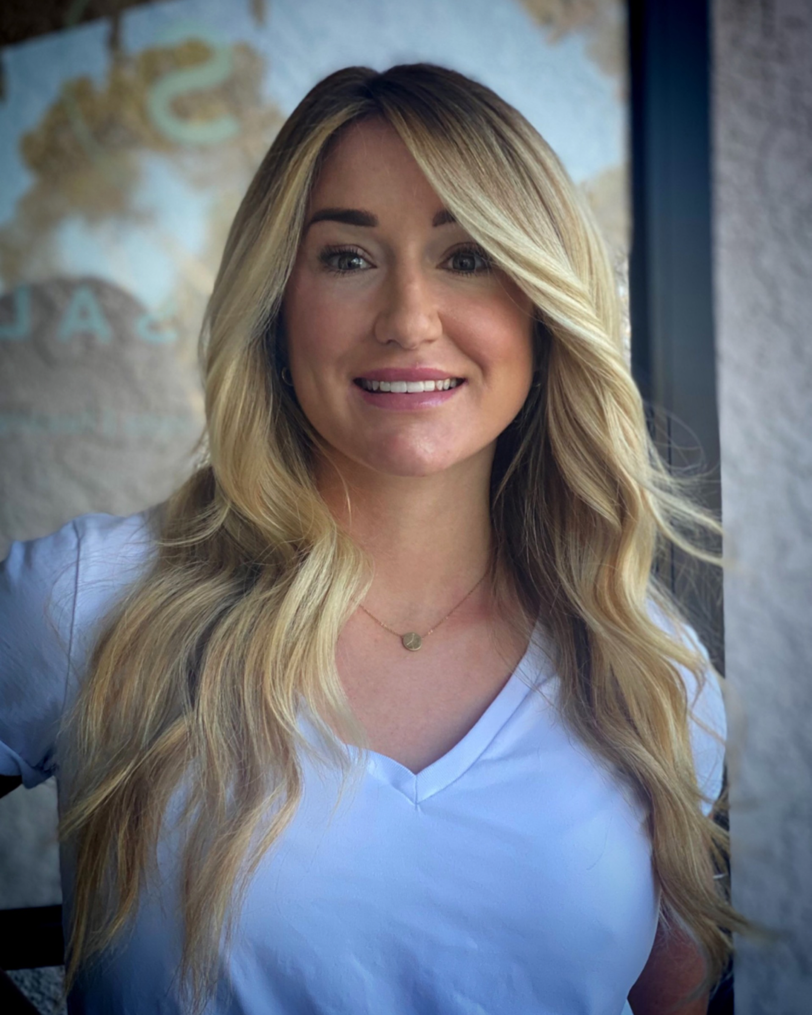 A woman with long, wavy blonde hair smiling, wearing a white top and a small gold necklace, standing indoors near a window with a partial view of a map or artwork in the background.