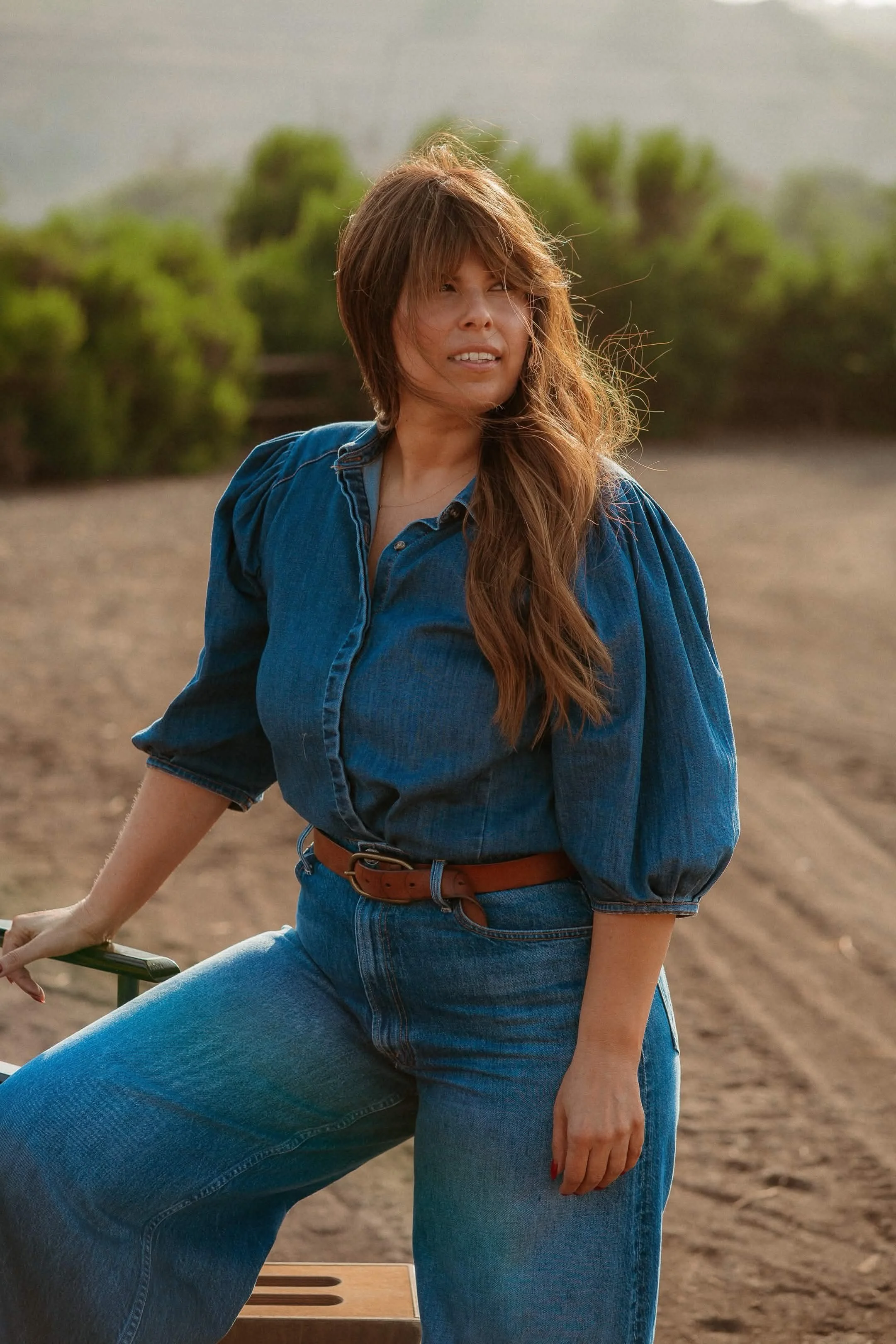 A woman with long wavy hair wearing a blue denim shirt and jeans, sitting outdoors on a farm or field with green bushes in the background, and twigs on her face.