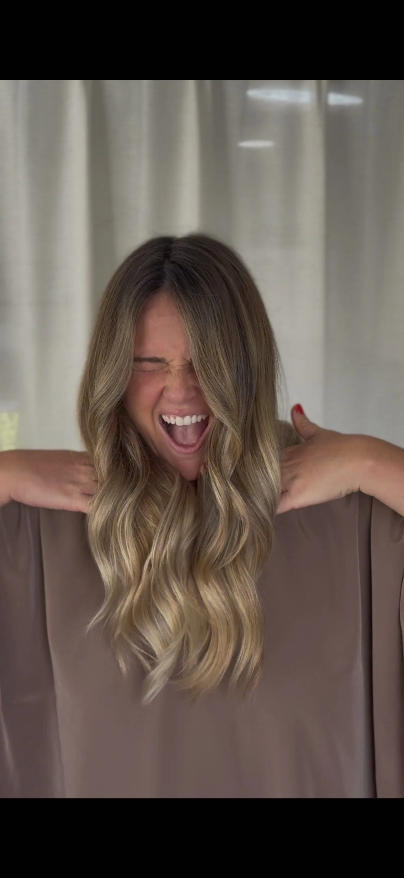 Woman with wavy blonde hair, mid-yawn or scream, holding her shoulders, standing in front of a curtain.