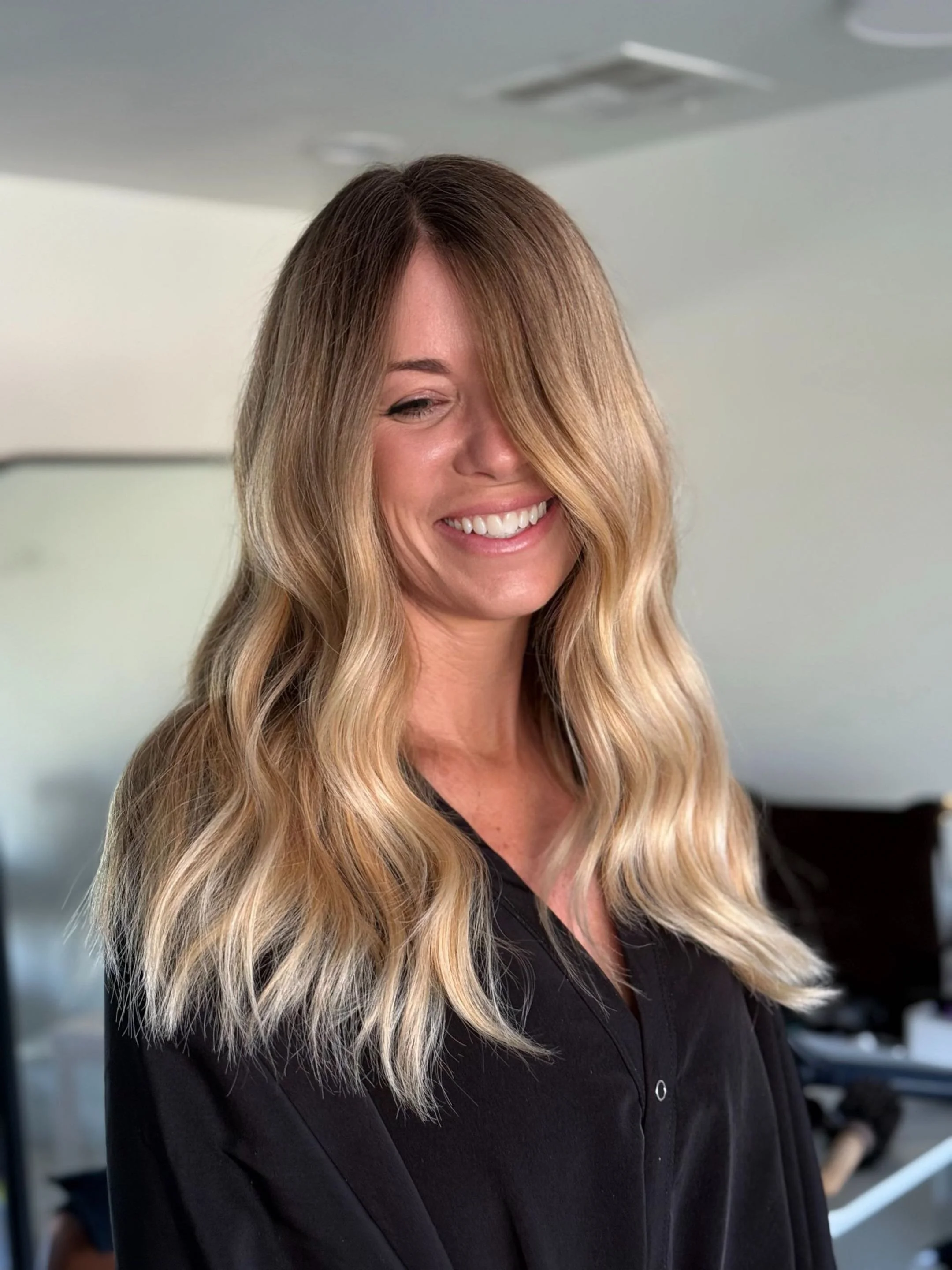 A woman with long, wavy, blonde hair smiling and looking downwards in an indoor setting.