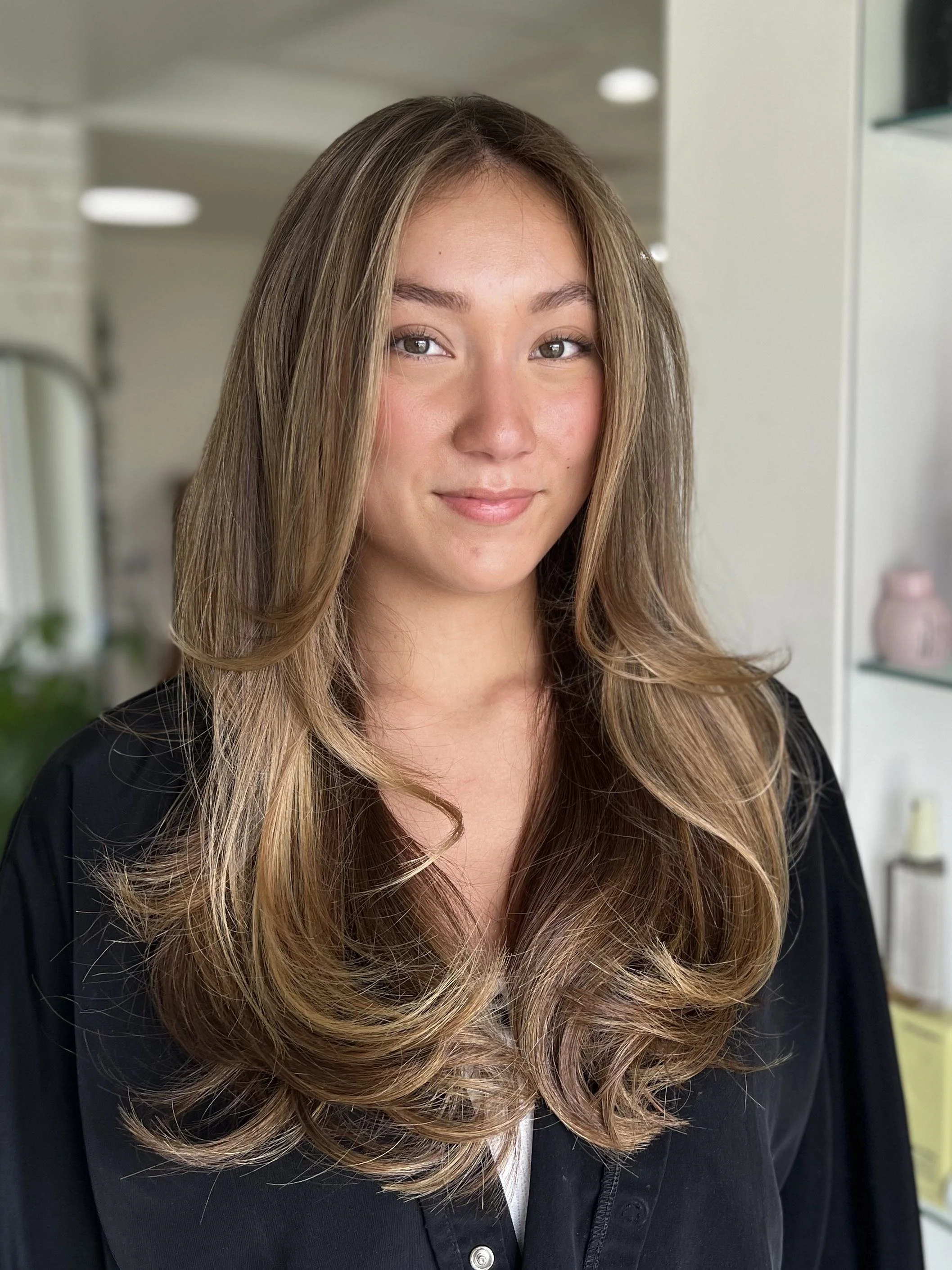 Close-up of a woman with long, wavy, light brown hair with highlights, wearing a black top, standing indoors with modern decor.