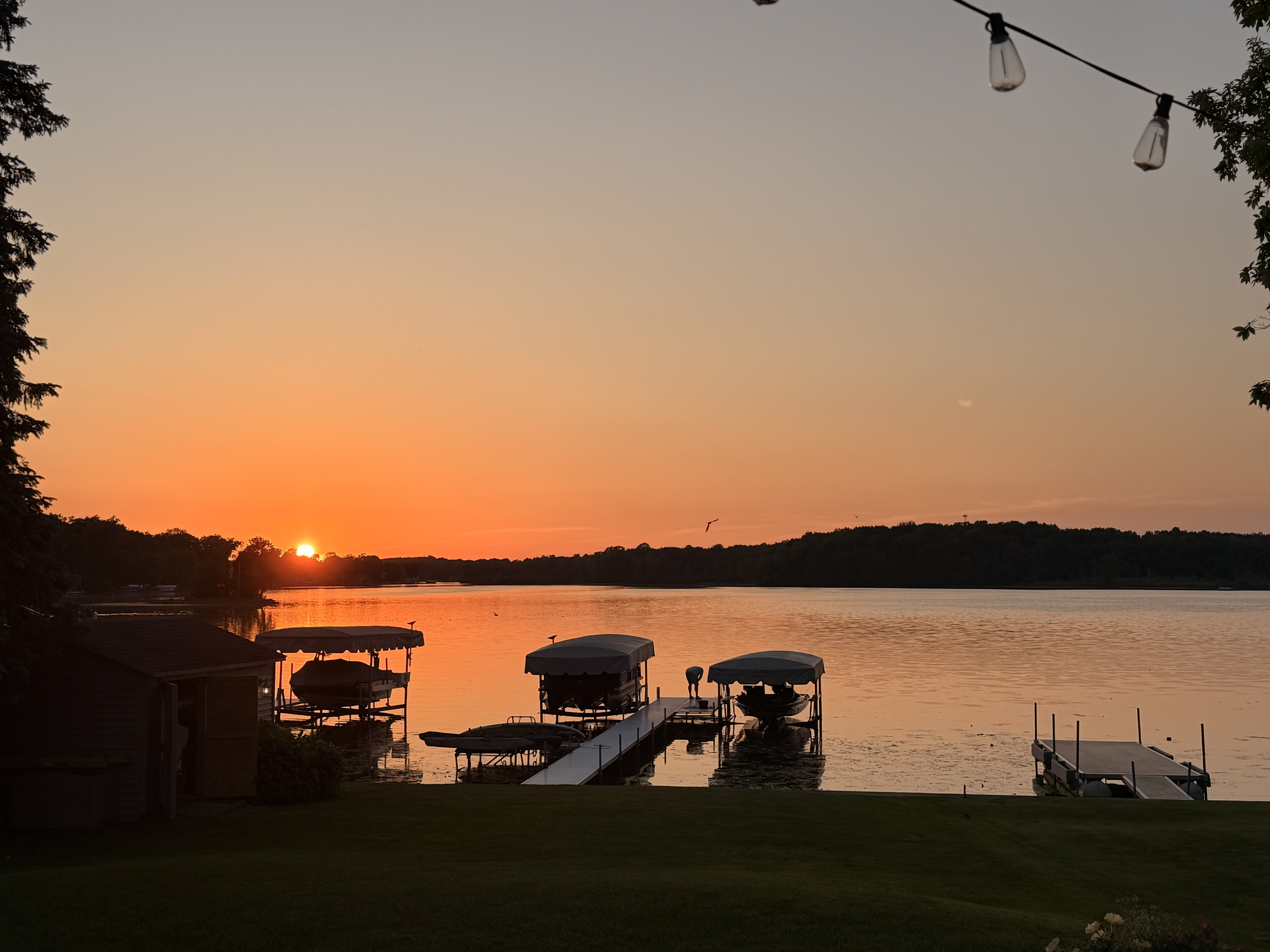 Sunset over a river or lake with boats at a dock, surrounded by trees and some buildings, with string lights hanging above.