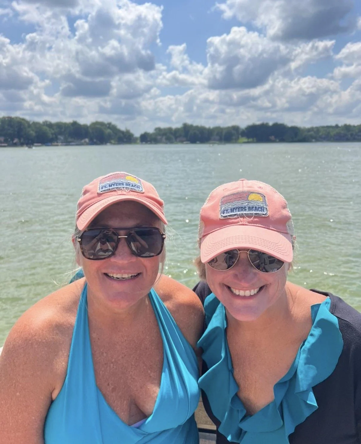 Two women wearing pink FUJI RIVERS BEACH hats and sunglasses, smiling, posing outdoors by a body of water under a partly cloudy sky.