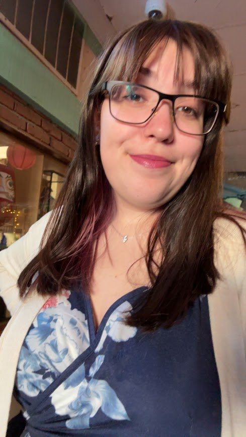 A woman with shoulder-length brown hair, wearing glasses, smiling, in a restaurant or cafe with brick walls and wooden decor.