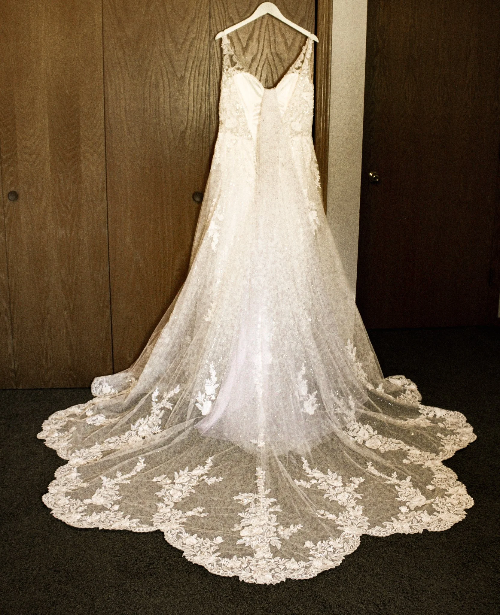A white wedding dress with lace detailing and a long train, hanging on a hanger against a wooden door.