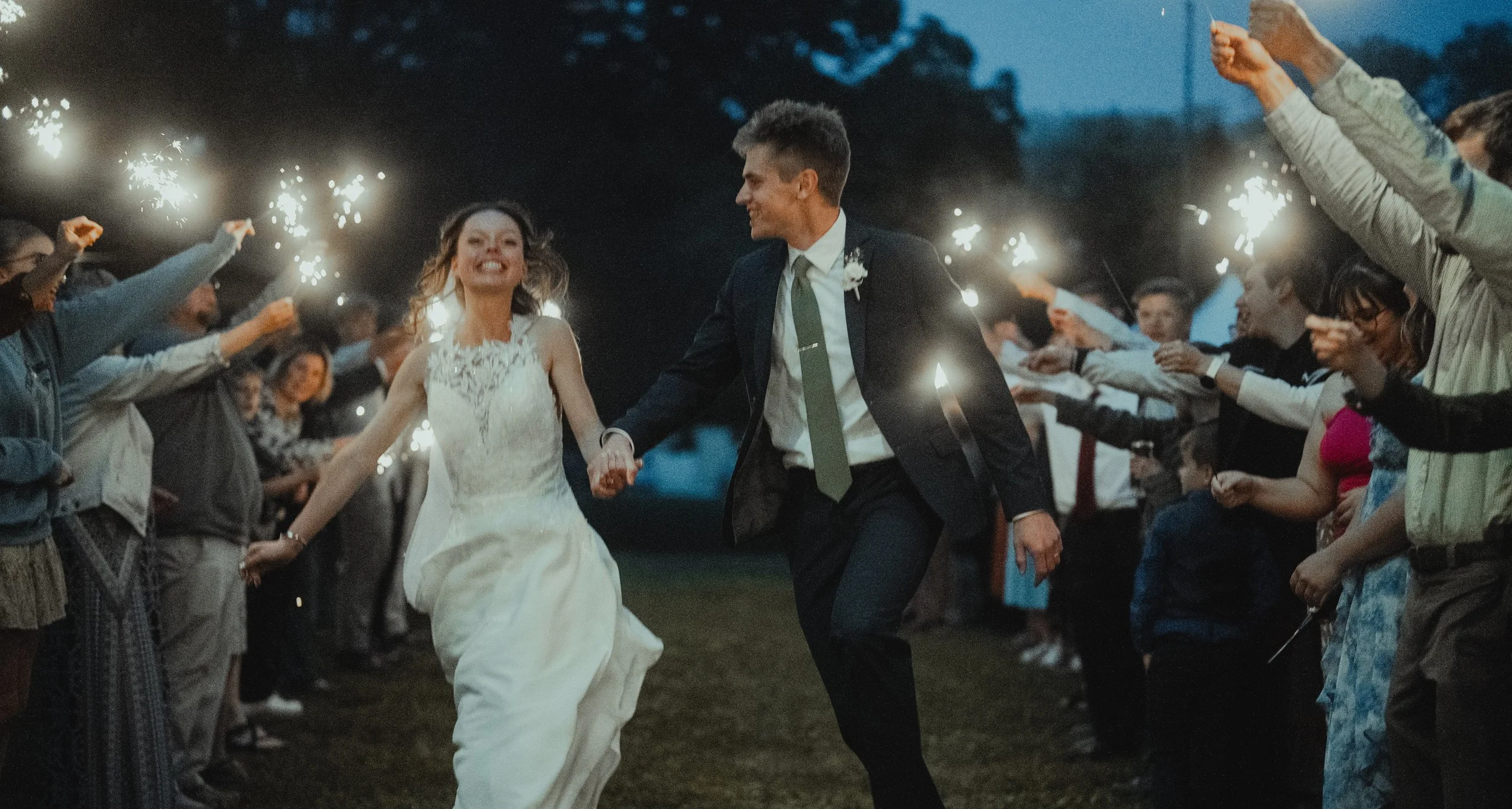 A newlywed couple walking hand in hand through a nighttime outdoor celebration, surrounded by guests holding sparklers.