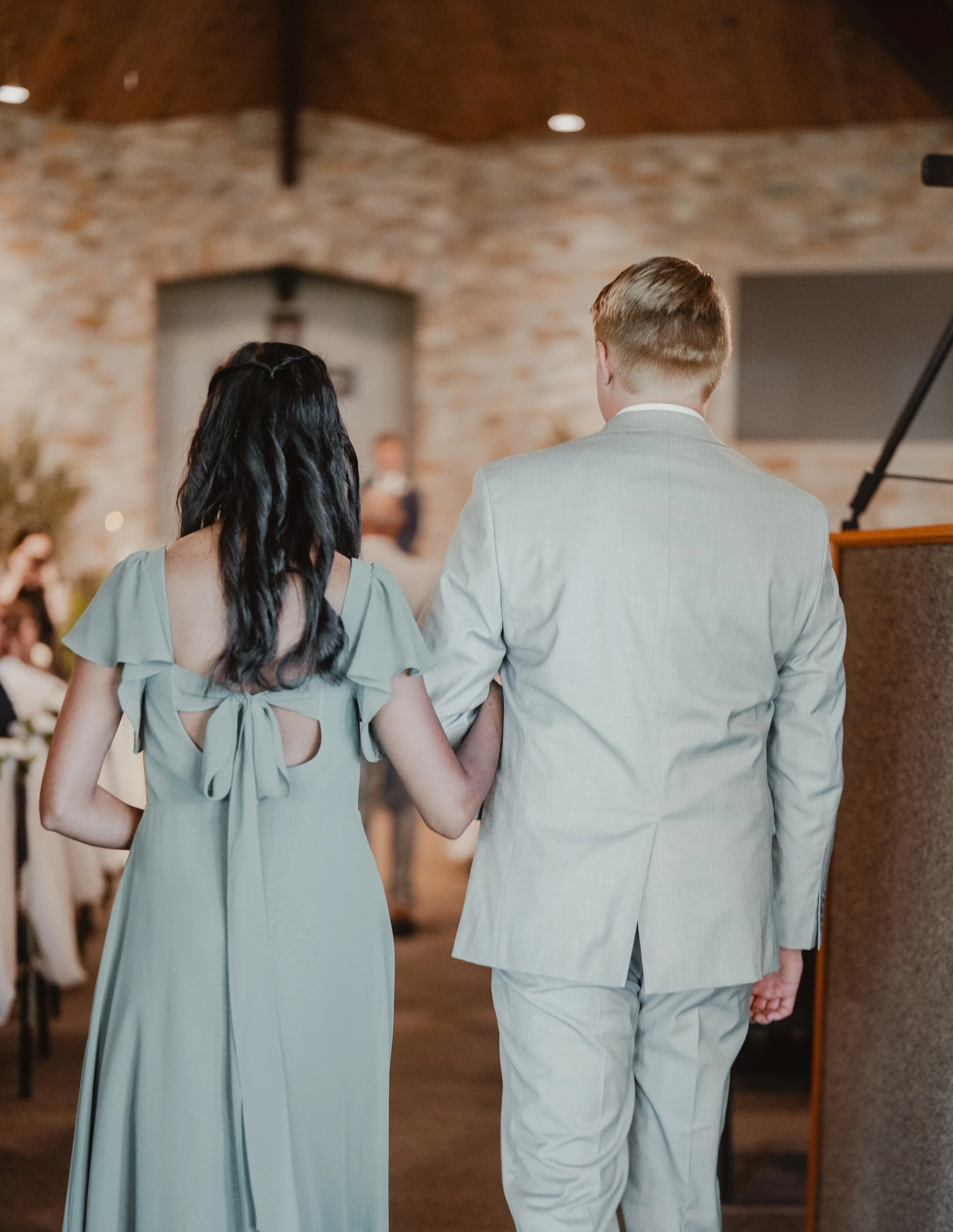A pair with arms interlocked and walking in a wedding or formal event setting, with a woman in a light green dress and a man in a gray suit, viewed from behind.