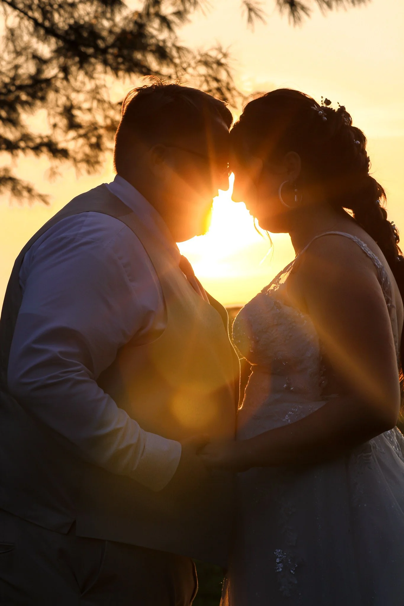 A silhouette of a couple in wedding attire leaning close together during sunset, with trees in the background, creating a romantic scene.