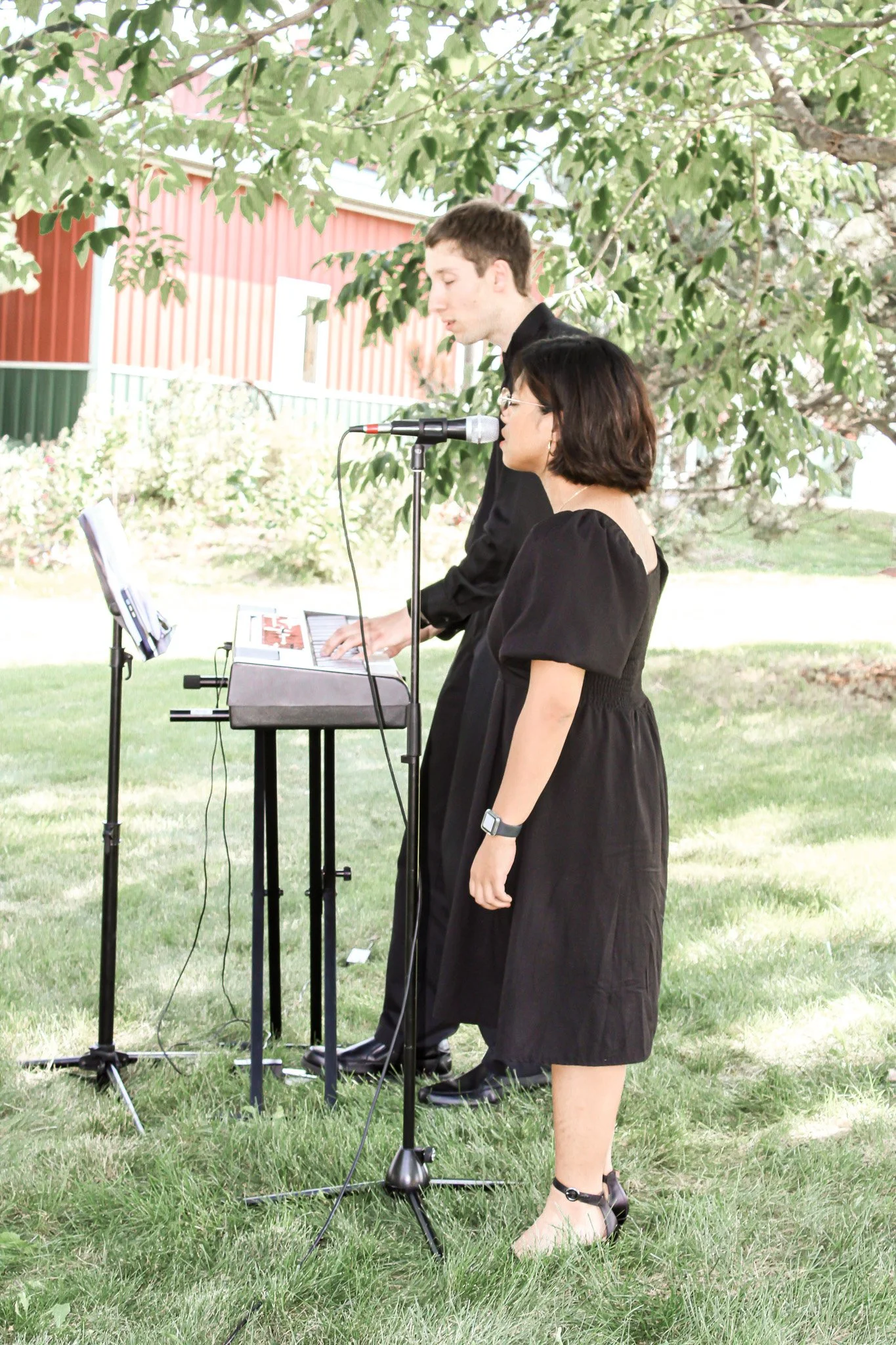 A young man and a woman are playing musical instruments outdoors under a tree, with a building with red siding in the background.