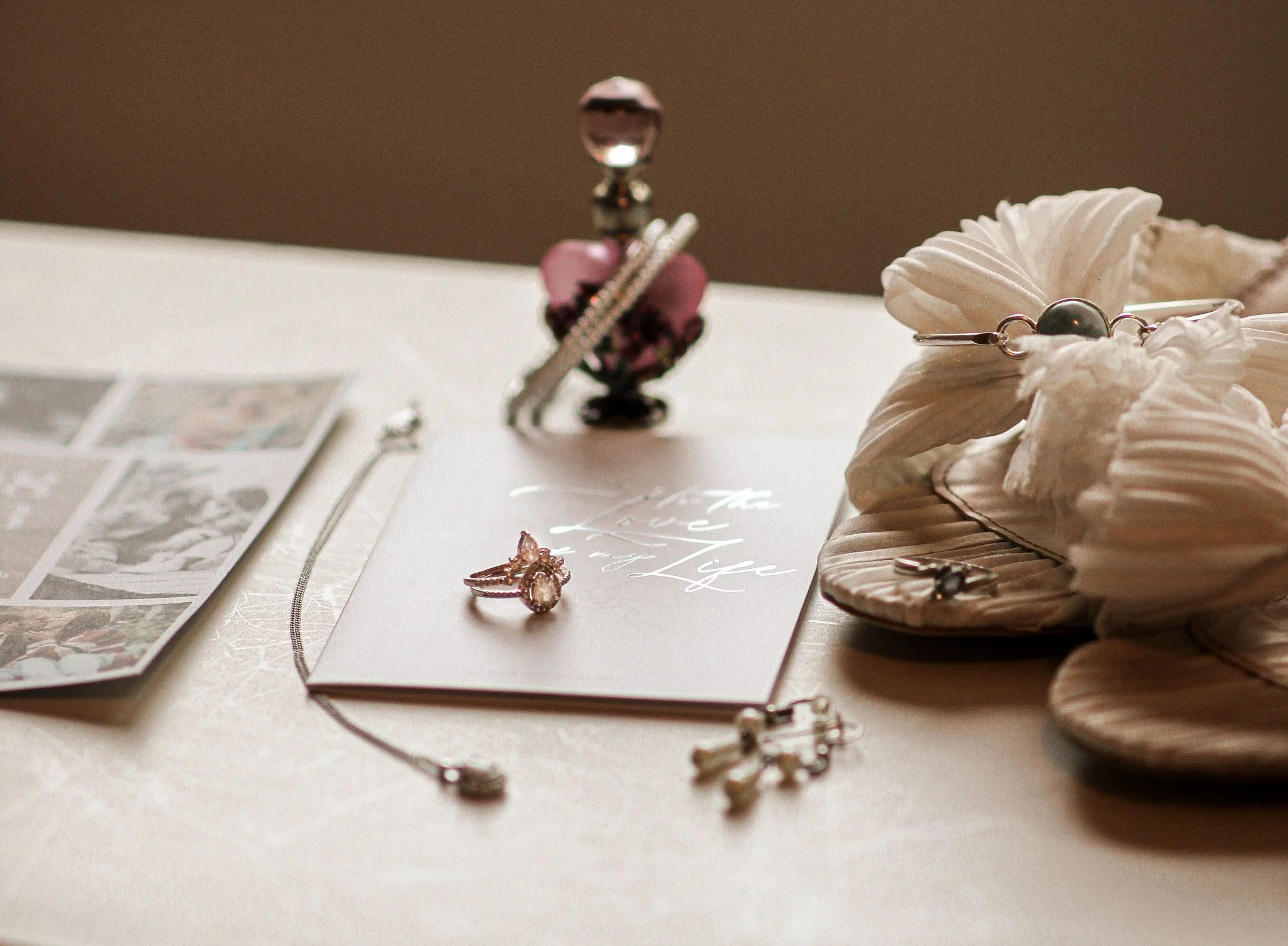 Jewelry display on a table with a ring, a bracelet, and a necklace, alongside wedding shoes, a white and silver booklet that reads 'to the love of my life,'  and a blurred image of an invitation.