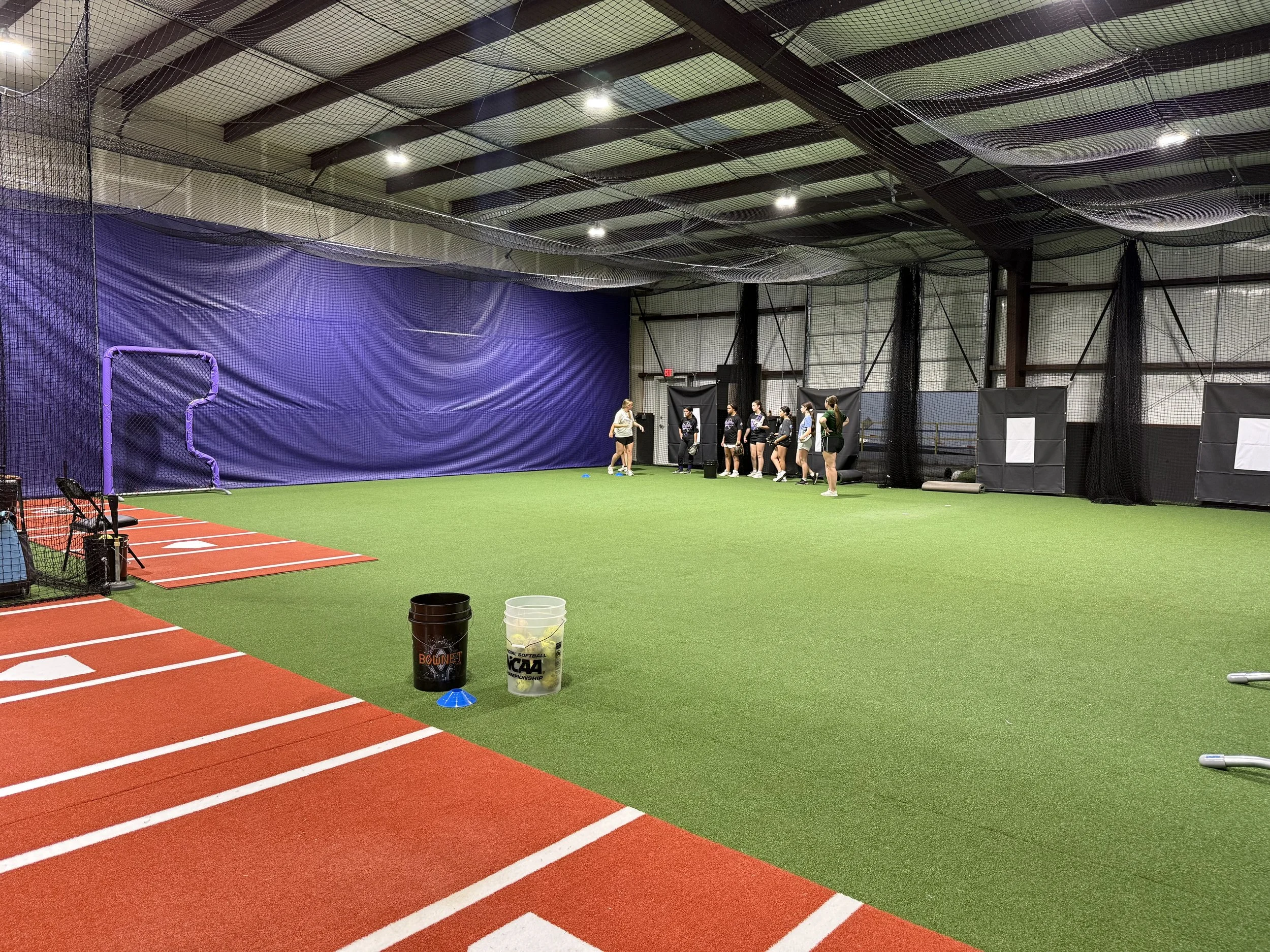 An indoor sports facility with a green turf, and black safety nets. There are several people standing in a line near the wall, possibly waiting for their turn or instructions. The ceiling has exposed beams and multiple lights.