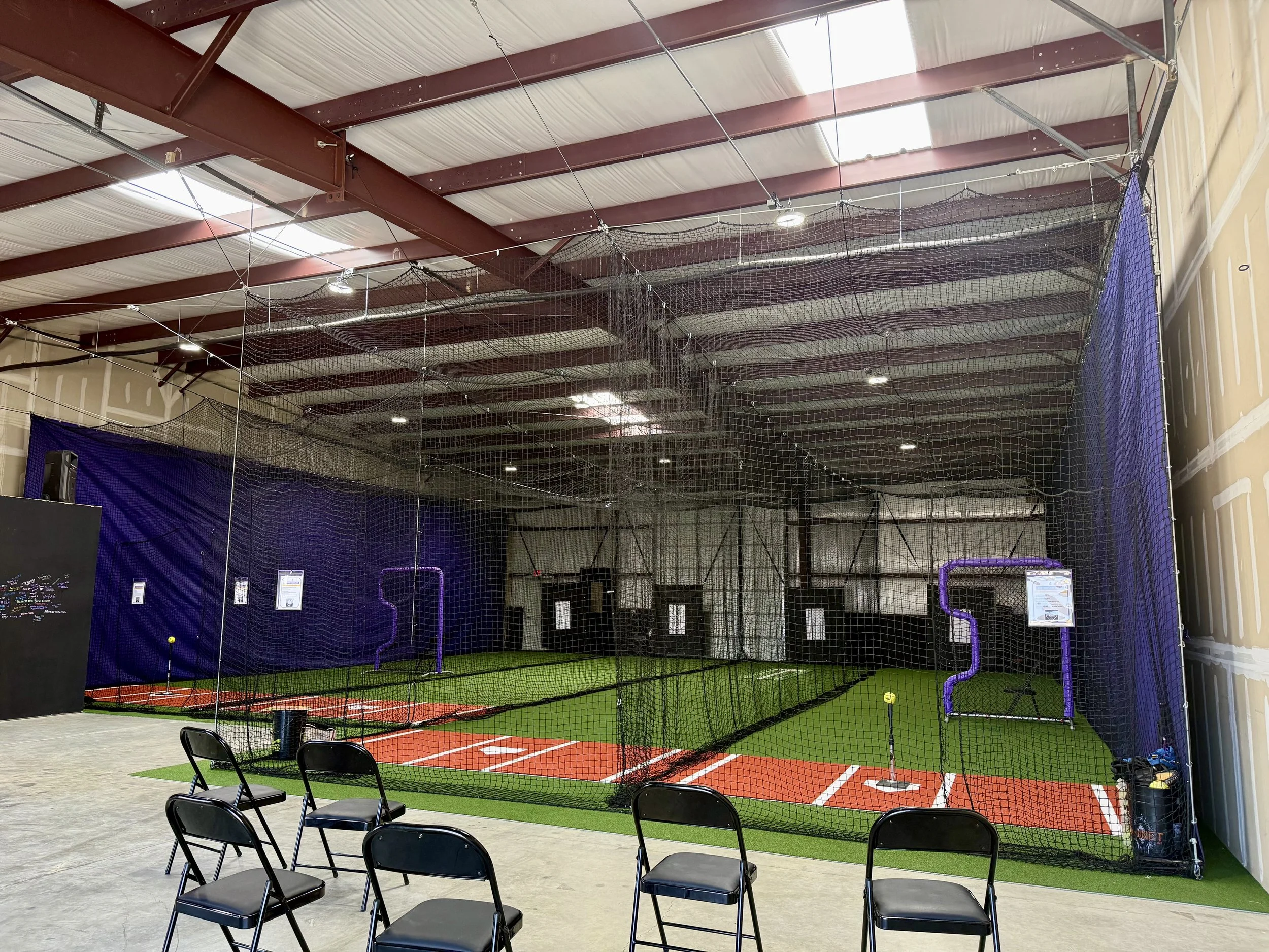Indoor baseball or softball training facility with artificial turf, pitching and batting cages, seating area with black chairs, and high ceiling with exposed red steel beams.