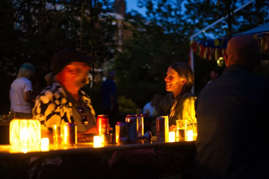 People gathered around a table with drinks and candles at an outdoor evening event on a summer evening.