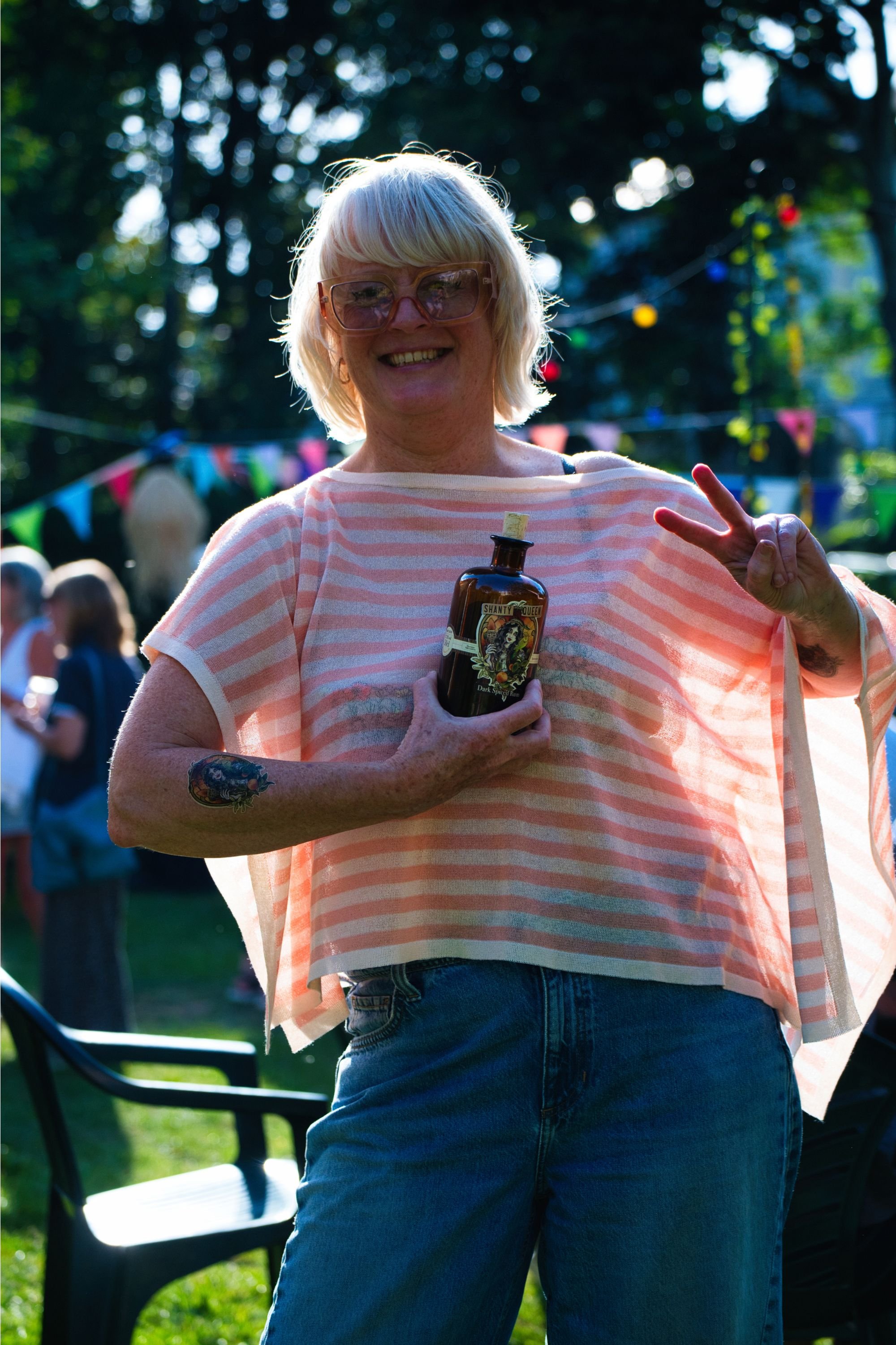 A woman with blonde hair, wearing large pink sunglasses and a striped top, smiling and making a peace sign at an outdoor gathering, a bottle of dark spiced rum in her hand and a tattoo of a woman's face on her arm.