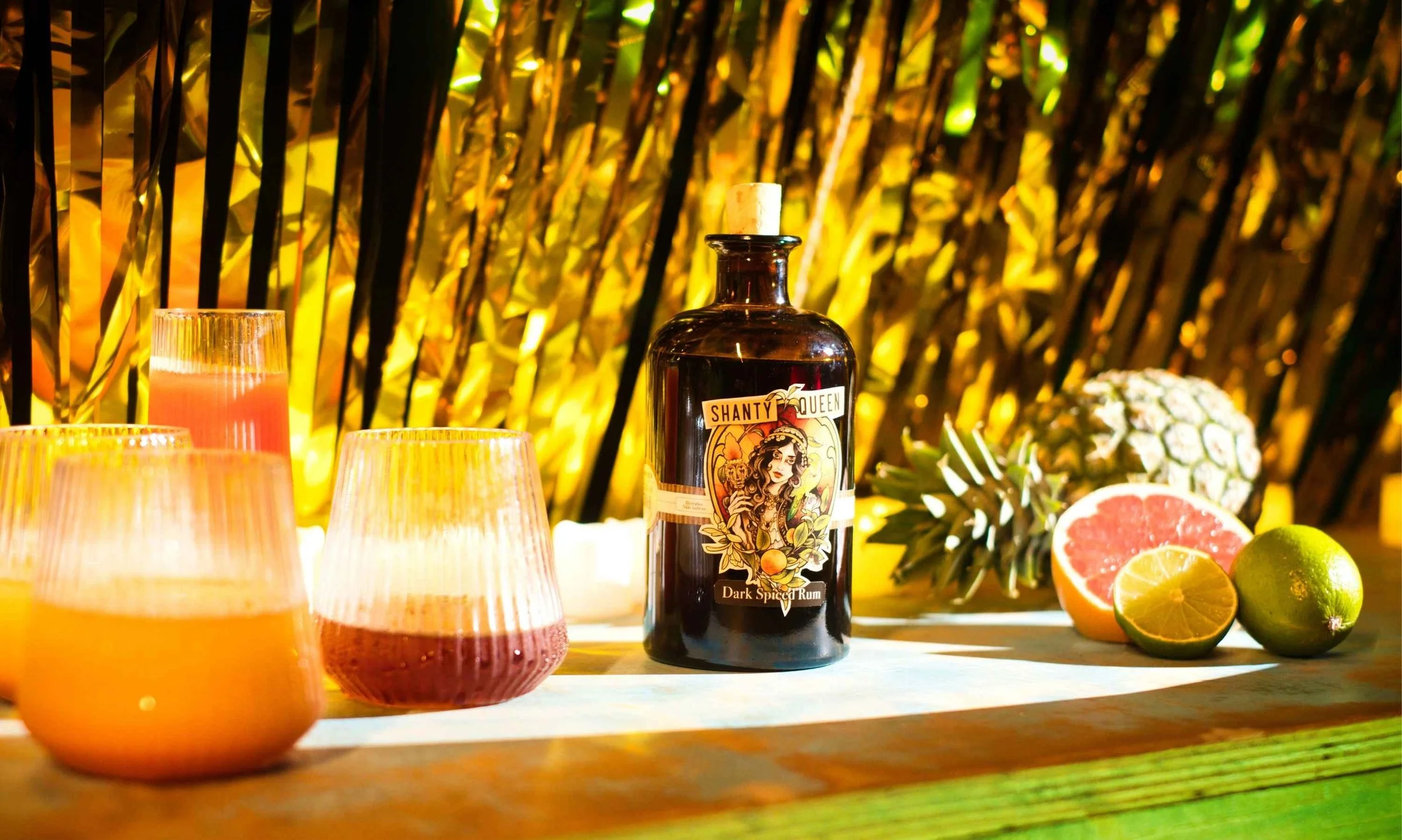 bottle of shanty queen spiced rum on a bar with cocktails in ribbed glasses on one side pineapple cut lime cut grapefruit on other gold shimmer curtains in background