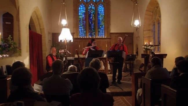 A live musical performance in a church with three musicians playing flute, bassoon and piano on a stage in front of an audience.