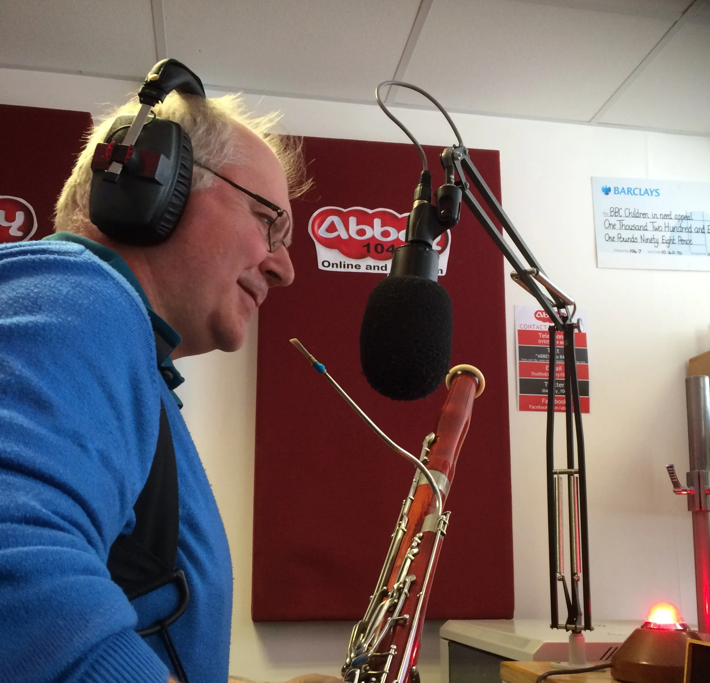A man with blonde hair and glasses wearing a blue fleece jacket, recording in a radio studio with a large microphone, headphones, and bassoon. The background has a red wall with the Abbey 104 logo and a white board.