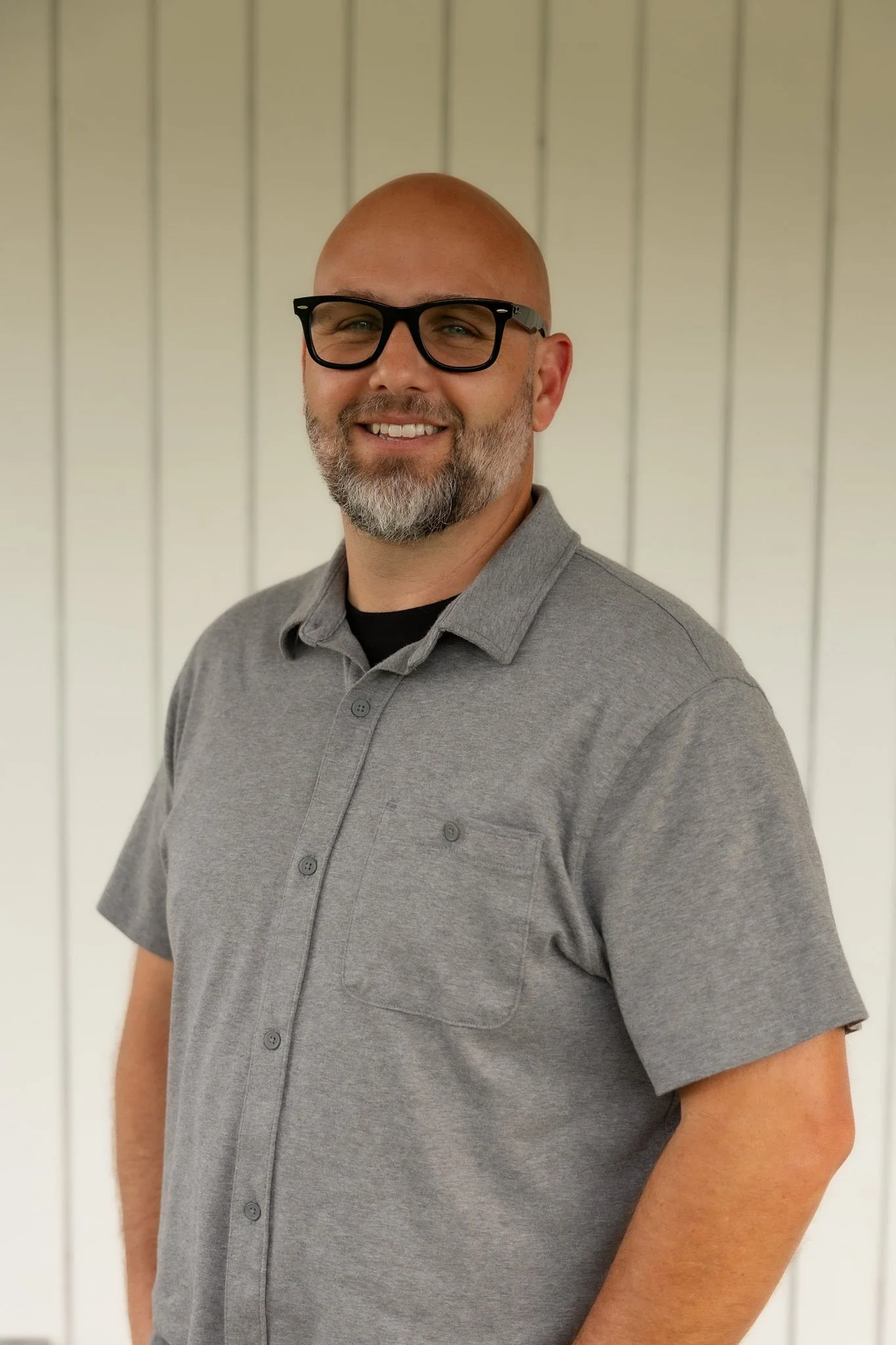 A smiling bald man with glasses and a beard, wearing a gray polo shirt, stands against a white wooden-paneled background.