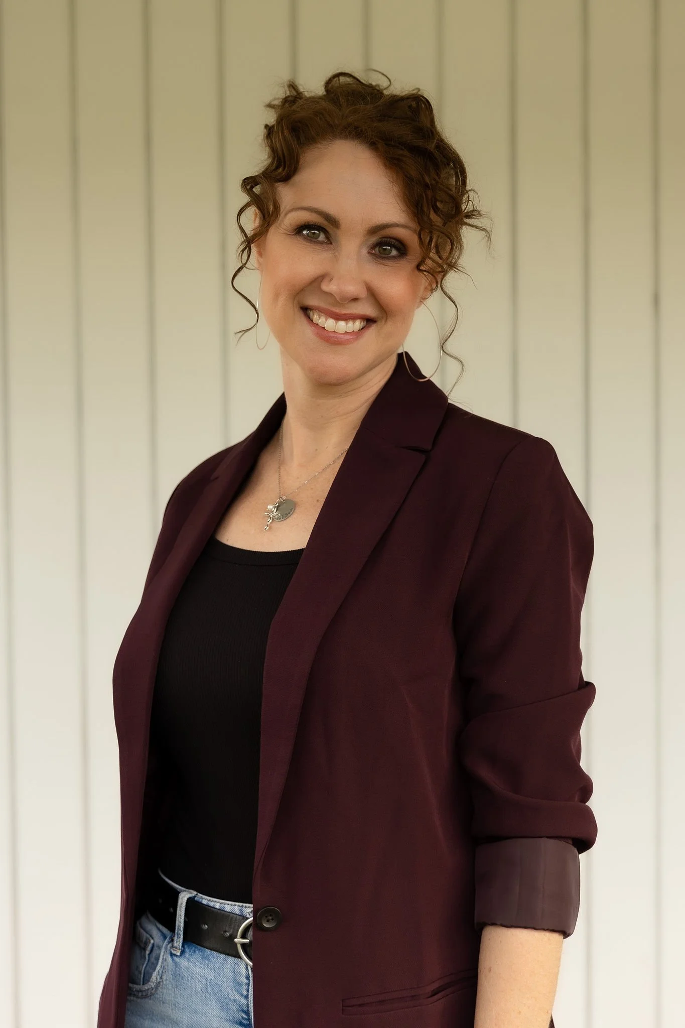 A woman with curly red hair wearing a maroon blazer, black top, and blue jeans, standing against a light-colored panel background, smiling at the camera.