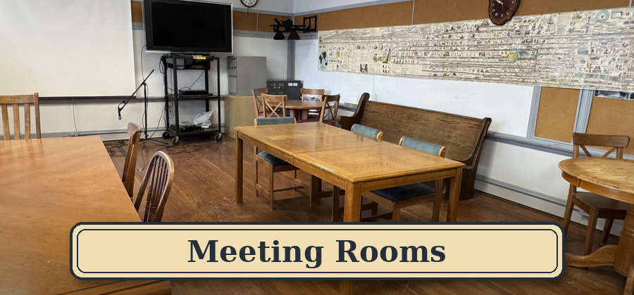 A classroom or meeting room with several wooden tables and chairs, a large wall map, a flat-screen TV, and a whiteboard. The room has hardwood flooring and a clock on the wall. There is a banner at the bottom with the text 'Meeting Rooms'. Blue Mount