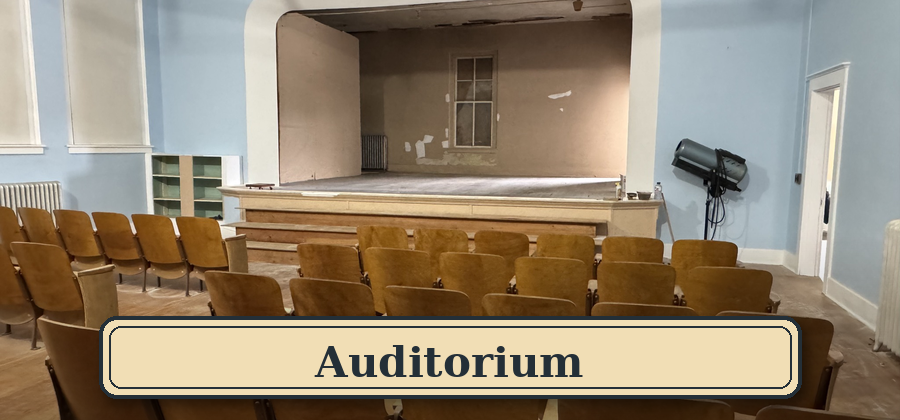 Empty auditorium with rows of brown seats facing a small stage with a plain wall and window at the back.