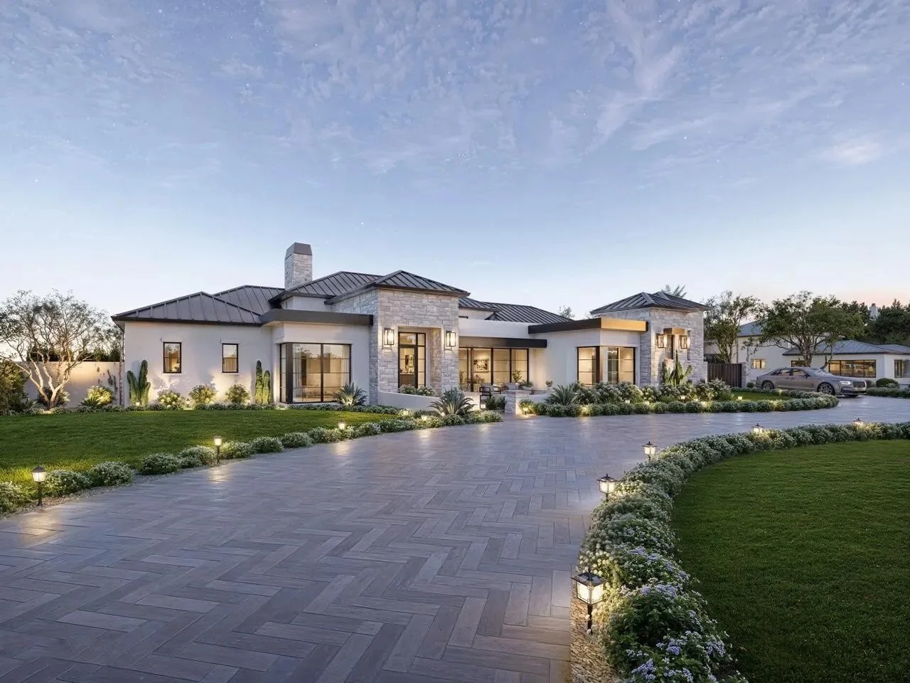 Modern single-story house with white walls, stone accents, large glass windows, and a dark metal roof. Front yard features a paved driveway, green lawn, and landscaped flower beds illuminated by small pathway lights, with trees and neighboring houses in the background under a clear sky.