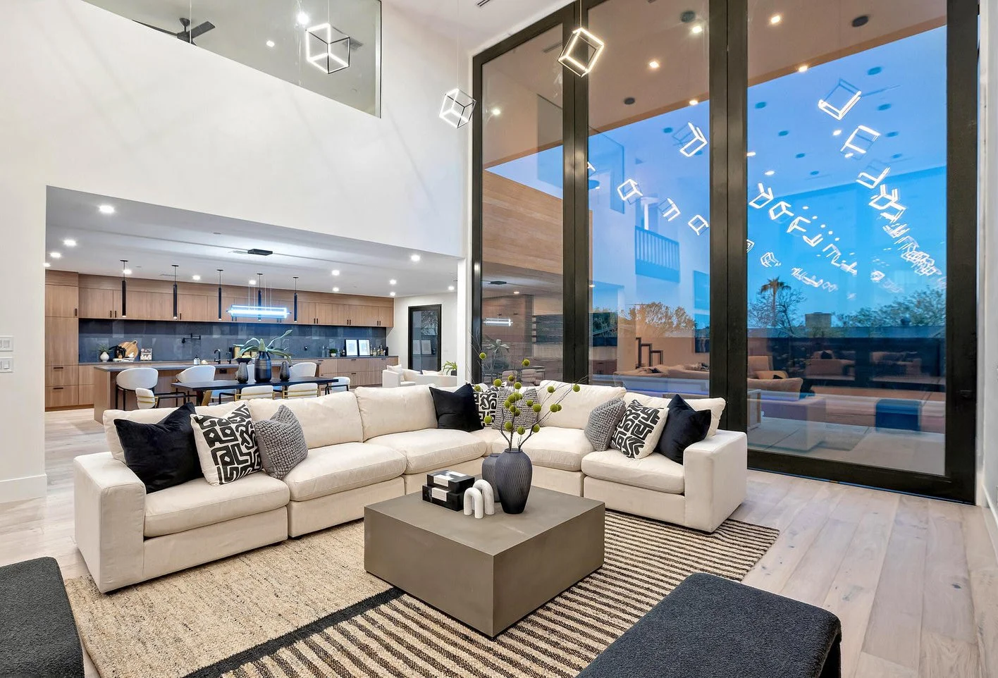 Modern living room with a large white sectional sofa, black and patterned throw pillows, a coffee table with decorative items, floor-to-ceiling windows reflecting hanging cube-shaped lights, a visible kitchen with wooden cabinets and a dining area in the background.