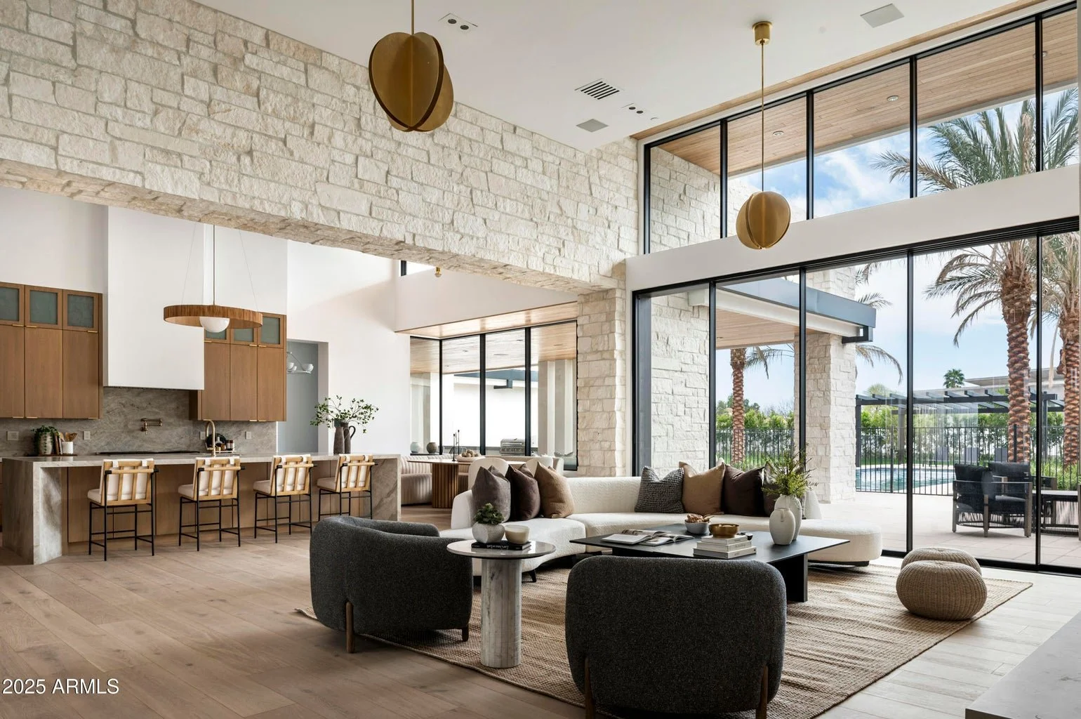 Modern living room with large glass windows, stone accent walls, a white sectional sofa, a black coffee table, and palm trees outside.