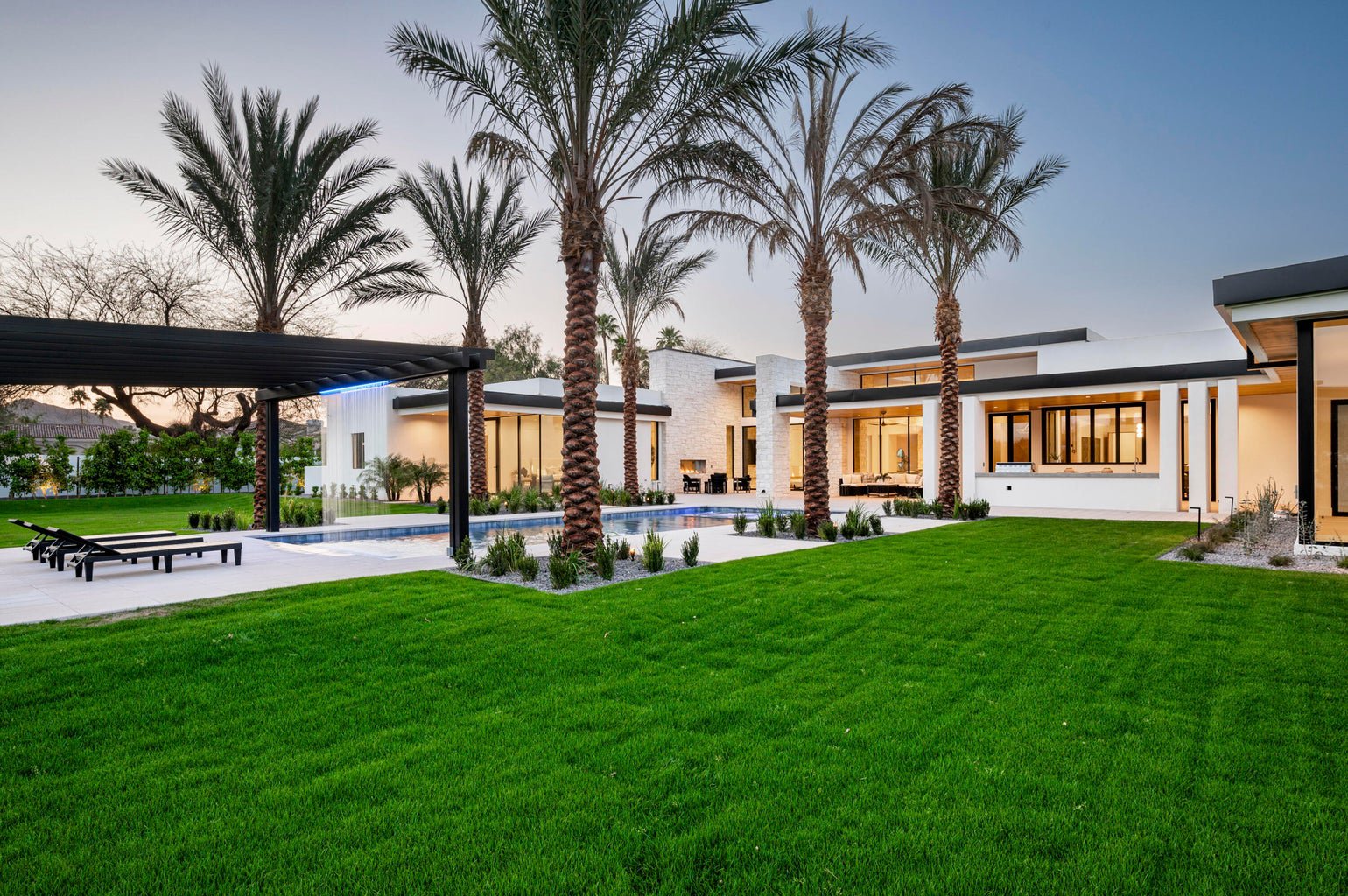 Modern house with well-maintained lawn and tall palm trees, outdoor pool, and patio area in the evening.