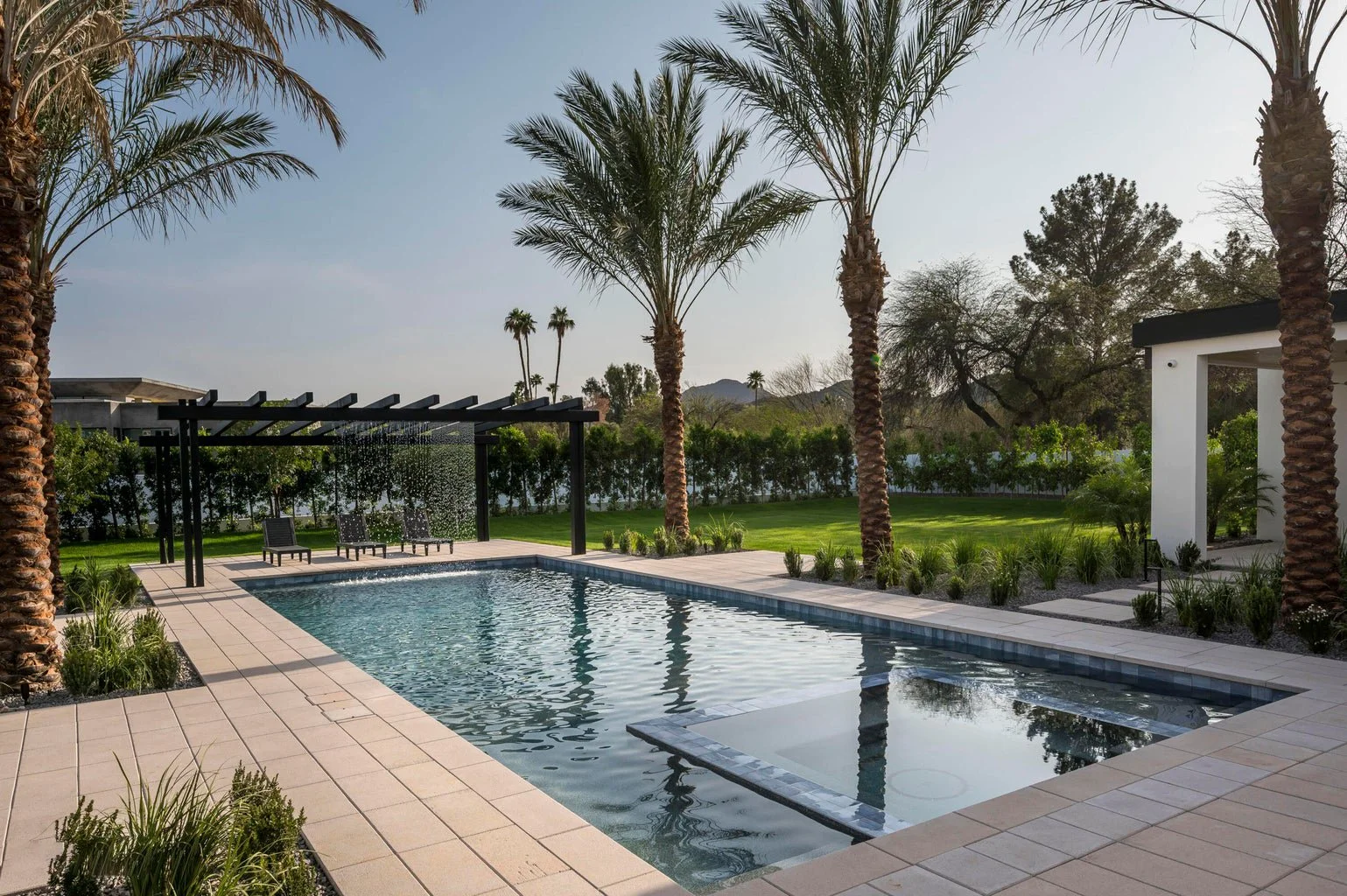 Sunset view of a modern backyard with a rectangular swimming pool, surrounded by palm trees, green grass, and a white building with a patio on the right. There are four lounge chairs under a pergola near the pool.