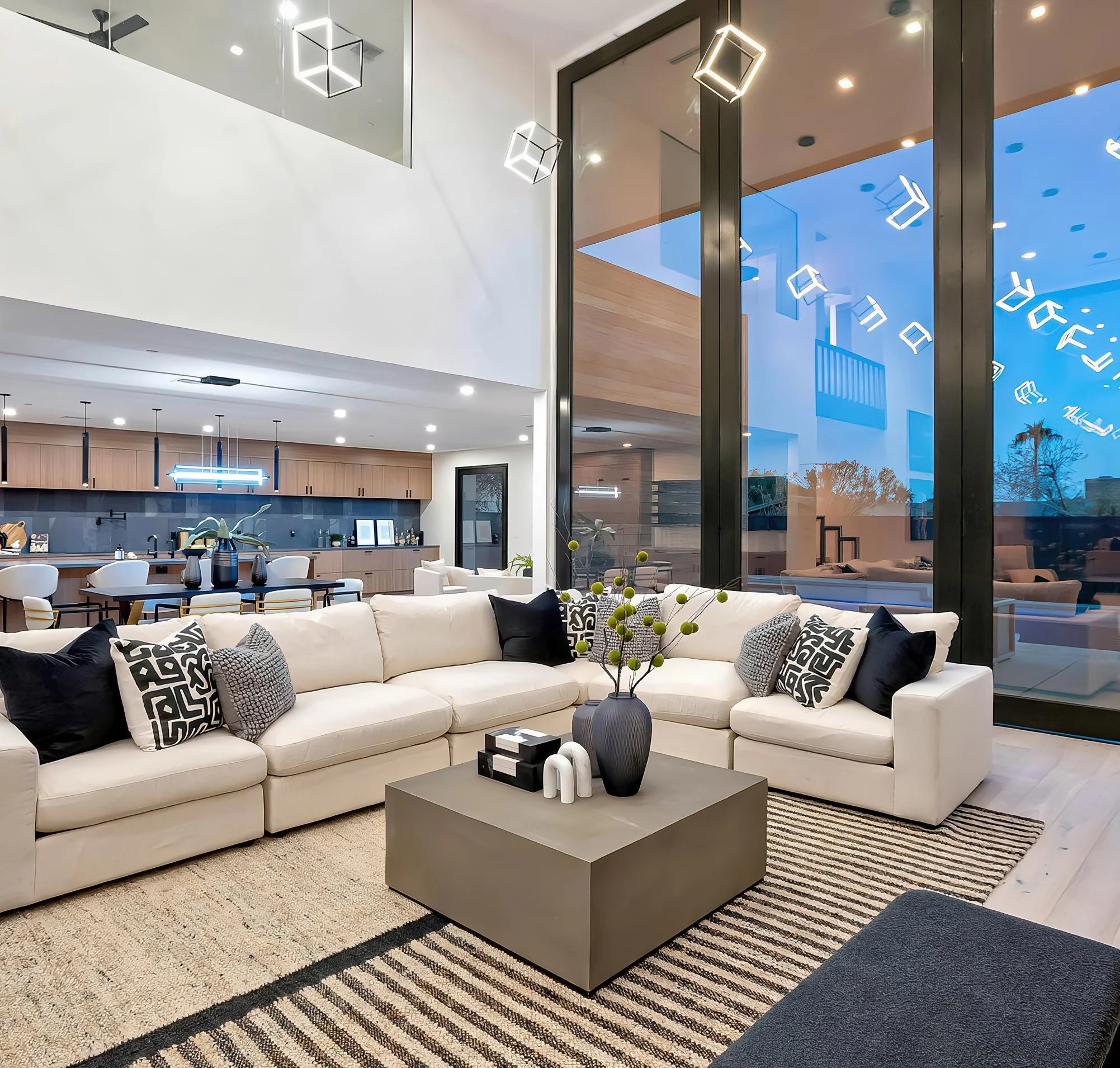 Modern living room with a large white sectional sofa, decorative black and white pillows, a dark coffee table, potted plants, a geometric area rug, large windows with reflections of contemporary light fixtures, and an open kitchen area with wooden cabinets.