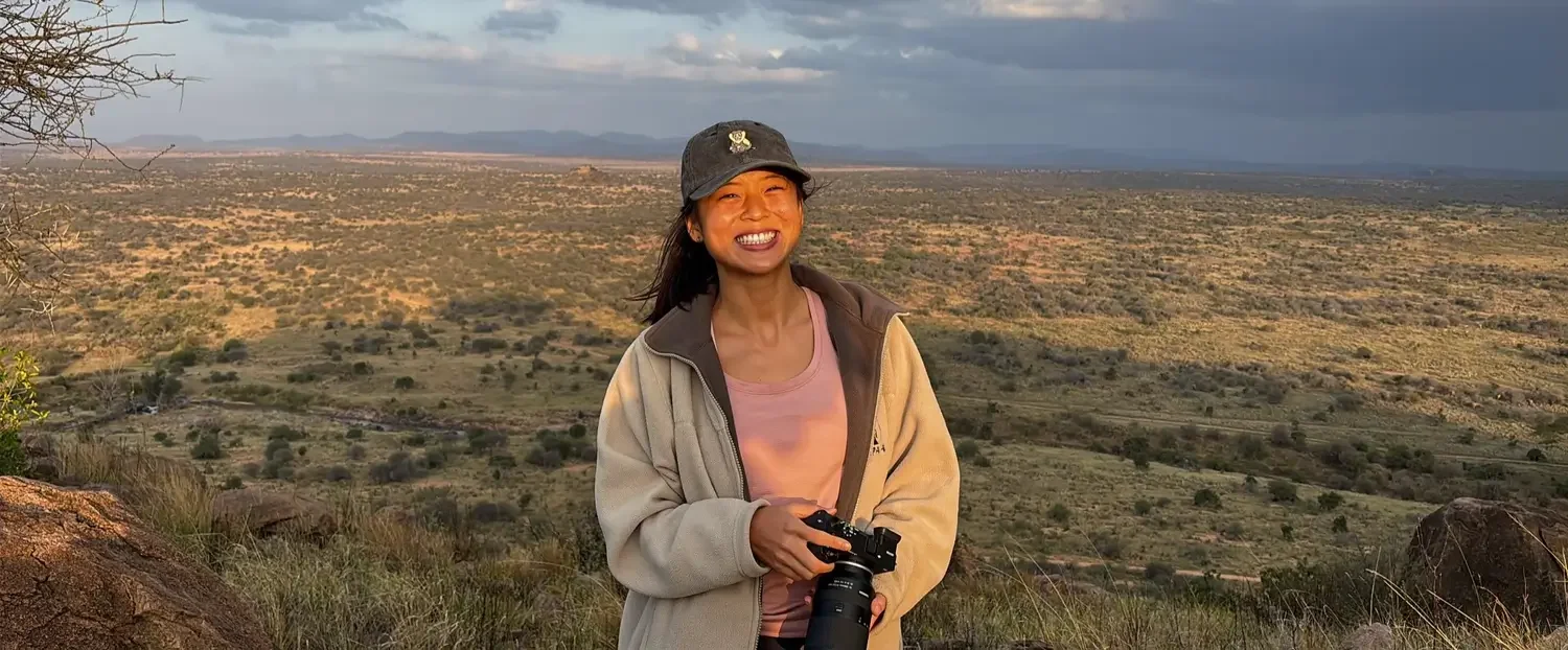 Eugenia smiling and holding a camera in front of a vast savannah landscape