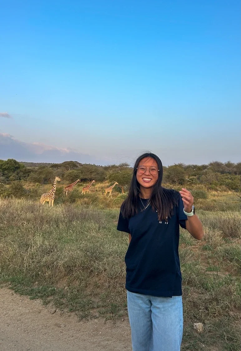 Eugenia smiling in the field in Kenya, with an open savanna landscape and wild giraffes behind her.