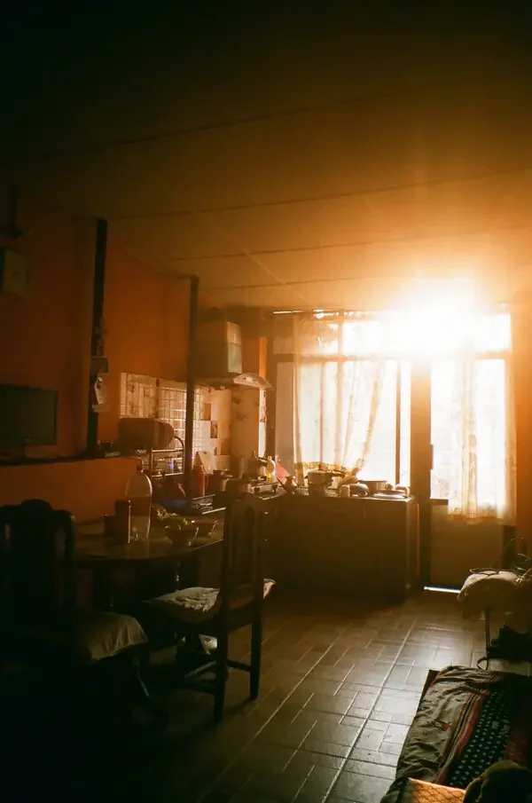 Bright, golden morning sunlight streaming through a large window into a dimly lit, cozy kitchen and dining area.