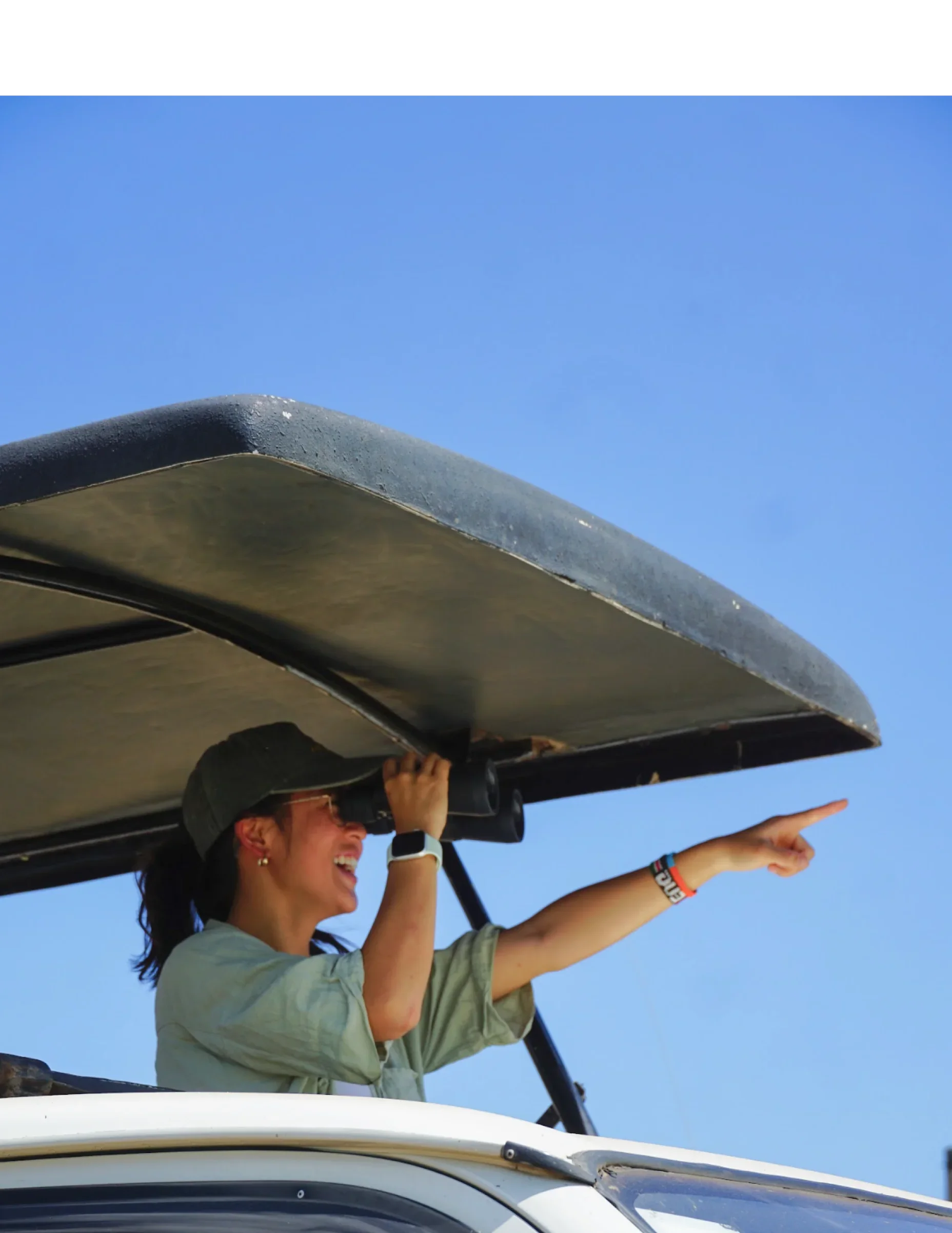 Eugenia looks through binoculars and points toward the horizon from the roof of a safari truck.