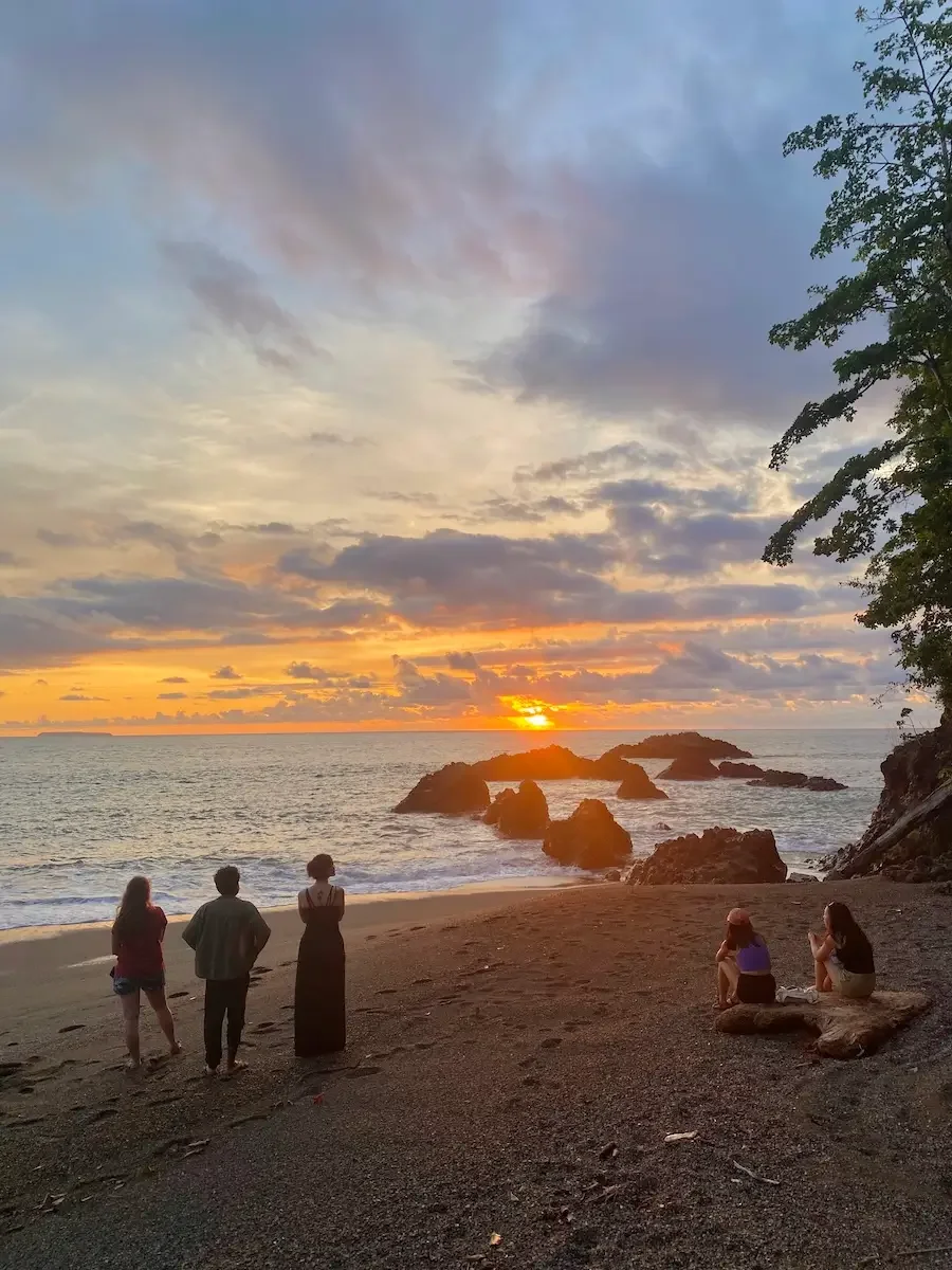 A group sits and stands on a dark sand beach watching a vibrant golden sunset over rocky ocean waves.