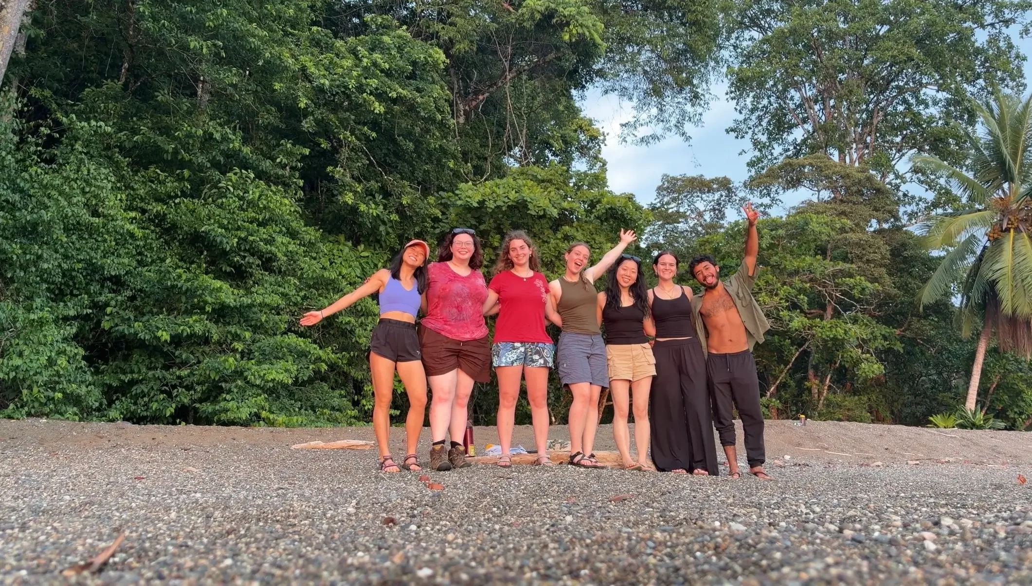 Seven travelers line up on a pebble beach, smiling and waving with a lush, green jungle wall directly behind them.