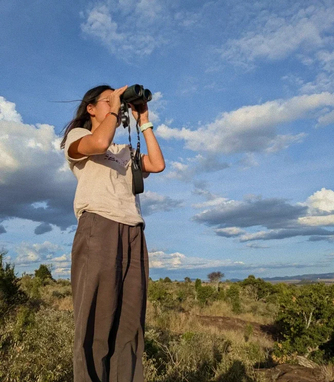 Eugenia looking through binoculars across a vast savannah landscape.