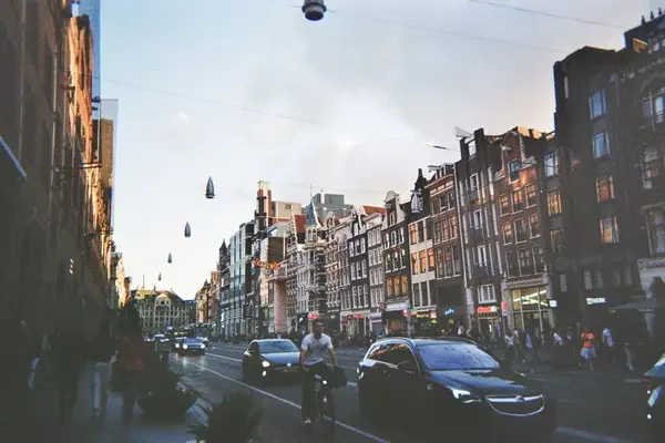 A cyclist commuting down a busy Amsterdam city street lined with tall, traditional brick buildings and passing cars at dusk.