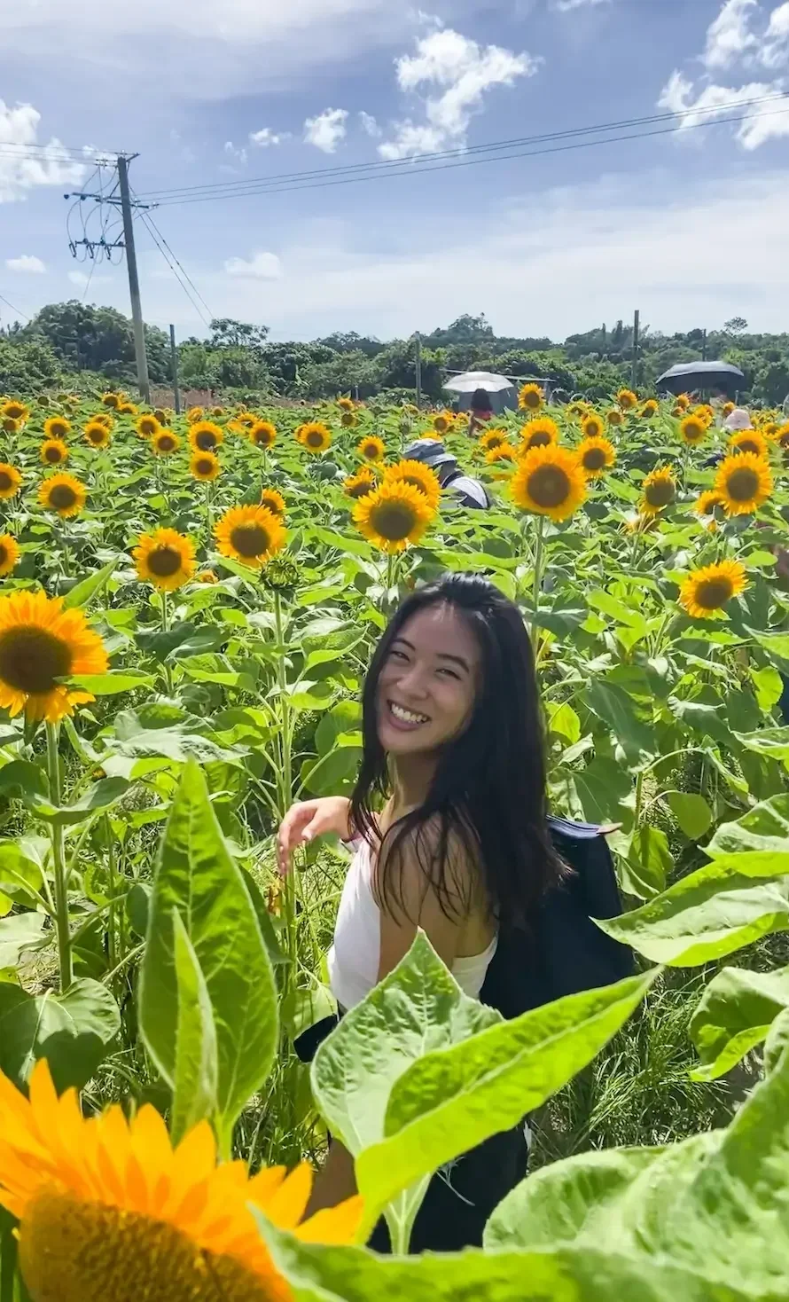Eugenia smiles warmly while walking through a vibrant field of tall blooming sunflowers.